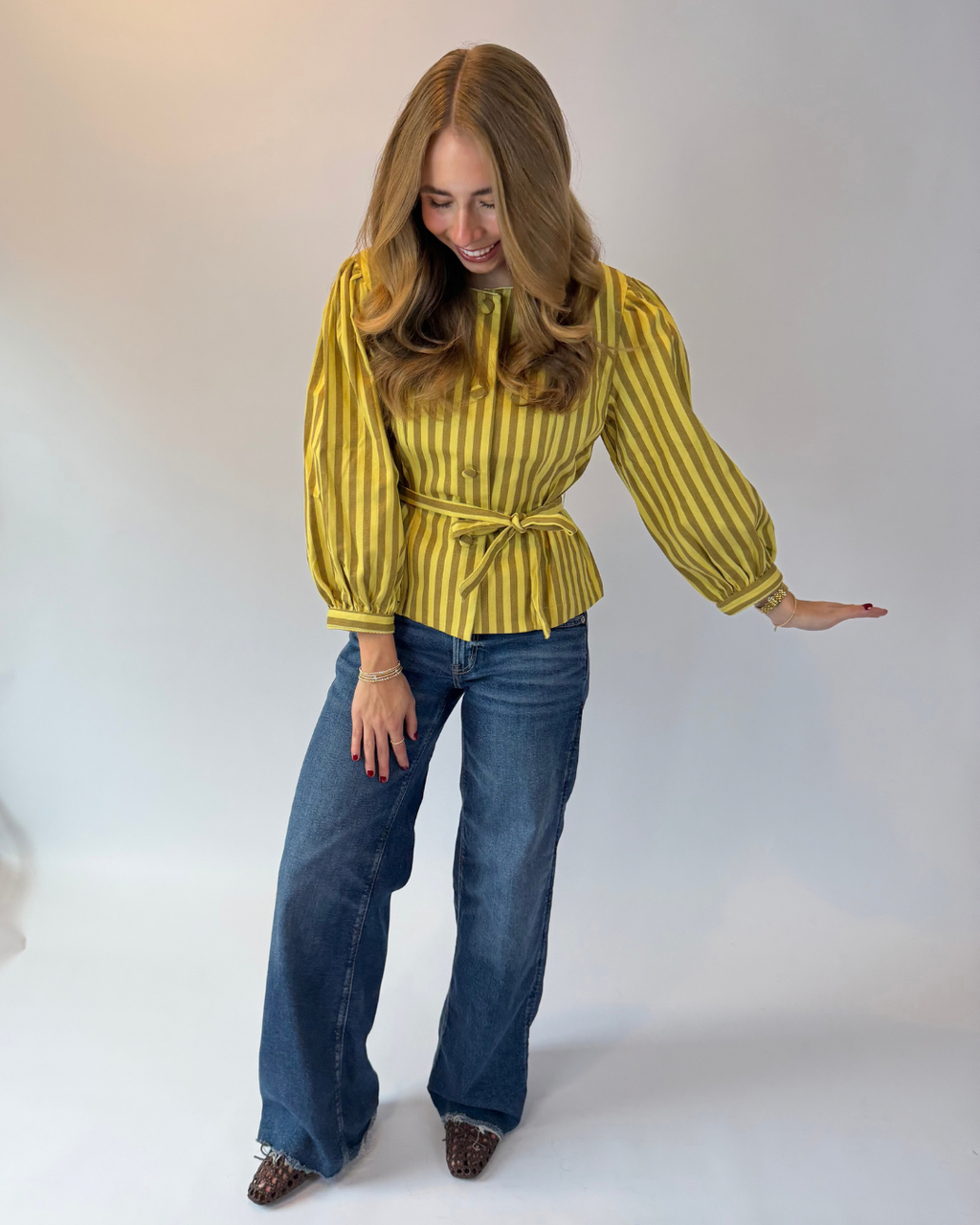 A woman modeling a yellow and dark green vertically striped long-sleeve blouse with a tie waist, paired with blue jeans and leopard print shoes, against a plain light gray studio background.