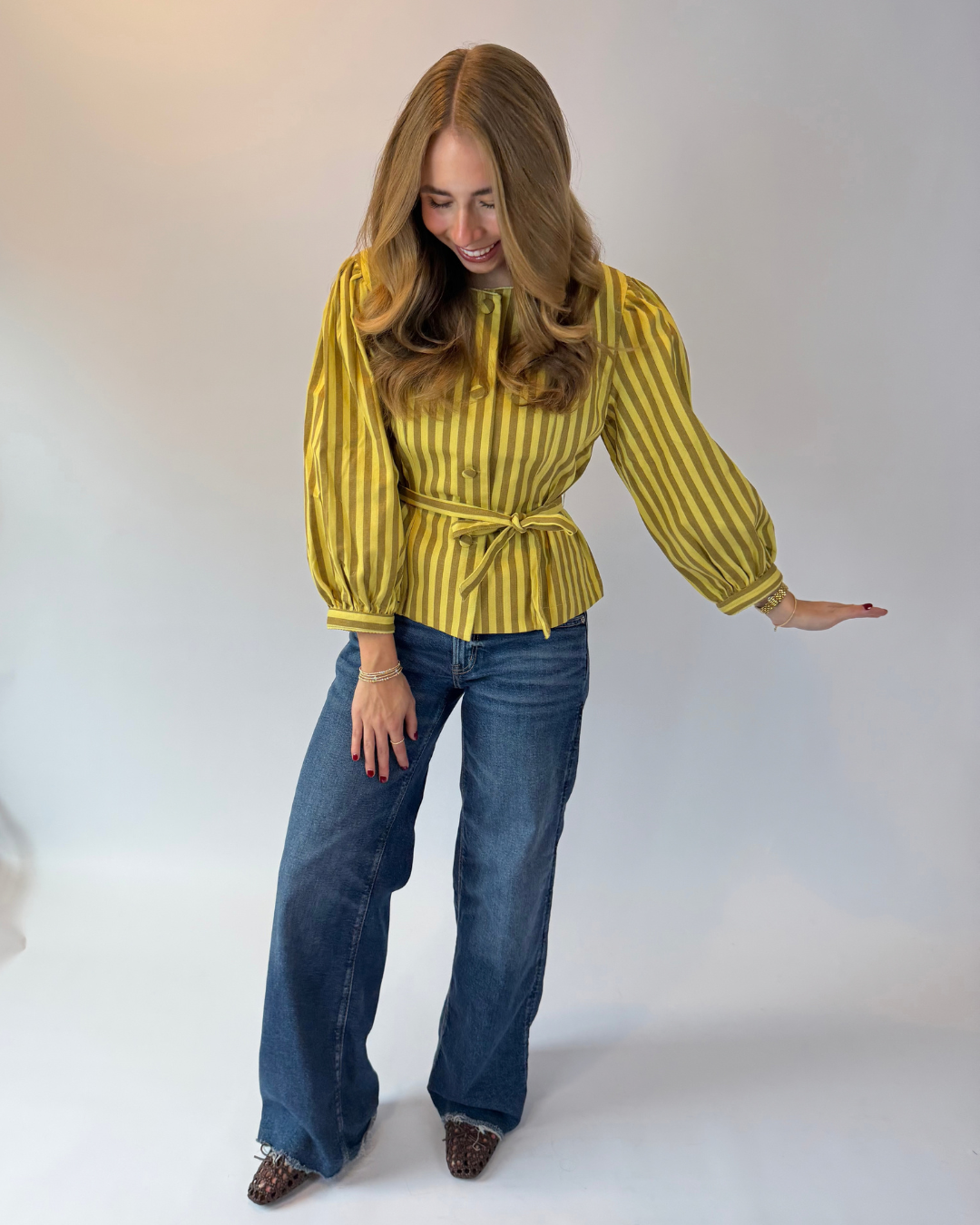 A woman modeling a yellow and dark green vertically striped long-sleeve blouse with a tie waist, paired with blue jeans and leopard print shoes, against a plain light gray studio background.