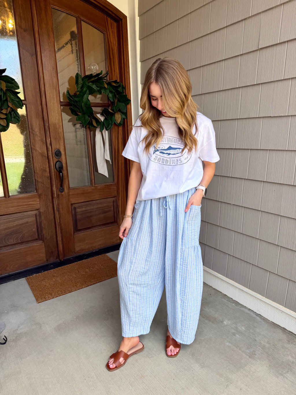Woman standing on a porch wearing a white t-shirt with a graphic design and light blue striped pants.