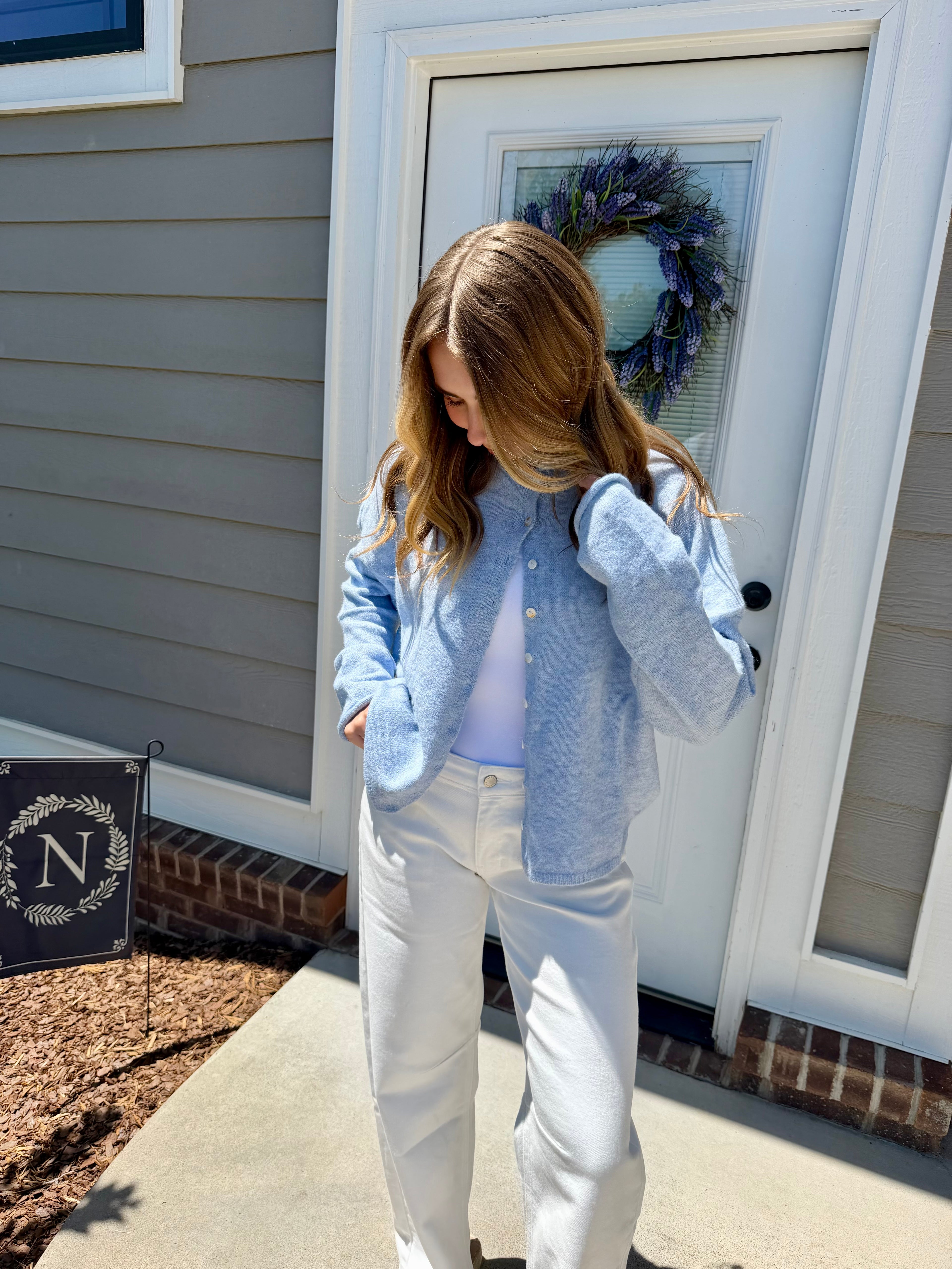 Woman wearing a light blue shirt and white pants standing in front of a house door with a wreath.