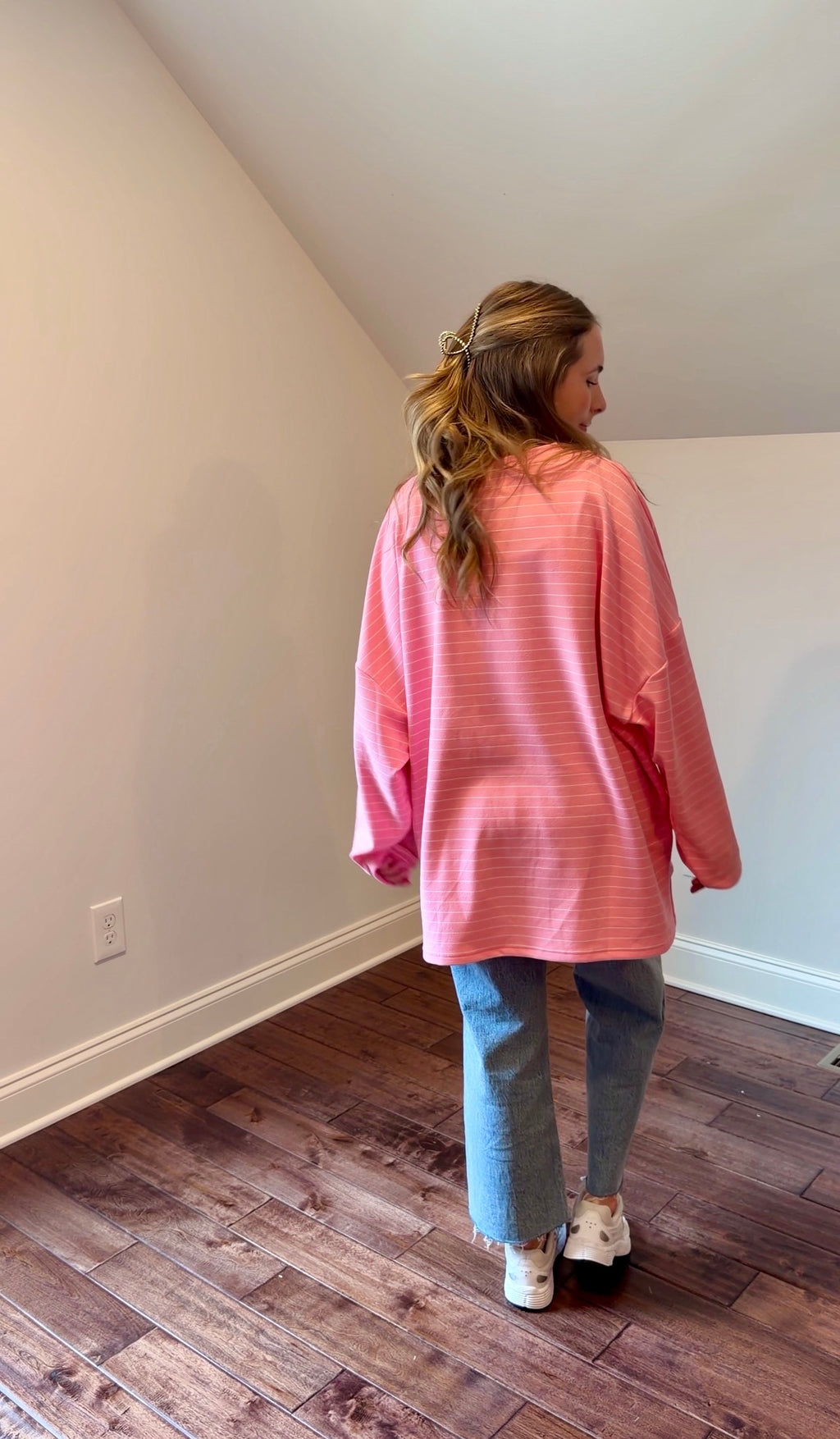 Person wearing a pink oversized shirt and blue jeans standing in a room with wooden flooring.