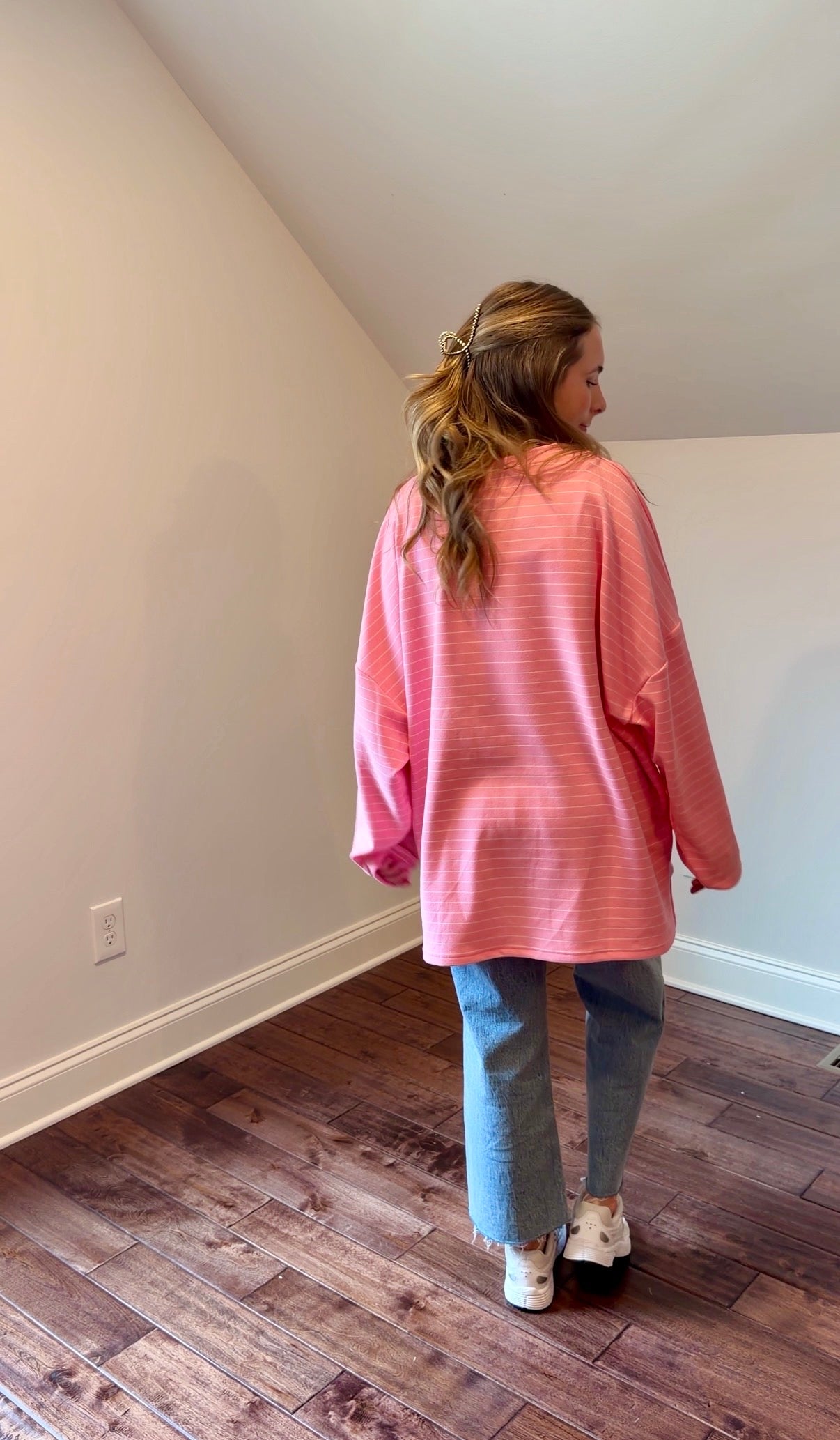 Person wearing a pink oversized shirt and blue jeans standing in a room with wooden flooring.