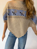 A woman wearing a beige knit poncho sweater with a light blue horizontal band featuring dark brown floral motifs and gray geometric patterns, paired with blue jeans.