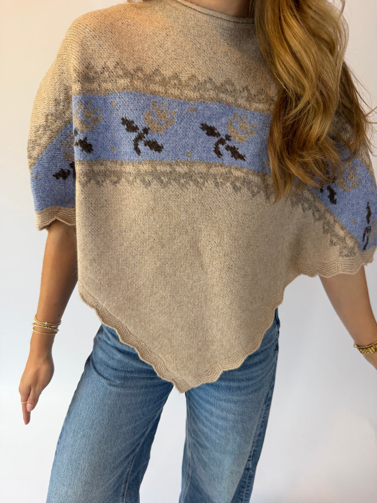 A woman wearing a beige knit poncho sweater with a light blue horizontal band featuring dark brown floral motifs and gray geometric patterns, paired with blue jeans.
