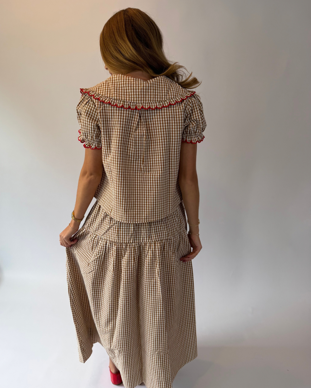 A woman viewed from behind wearing a beige and brown gingham two-piece set with red trim details, consisting of a short-sleeve top and a matching midi skirt, against a plain white studio background.