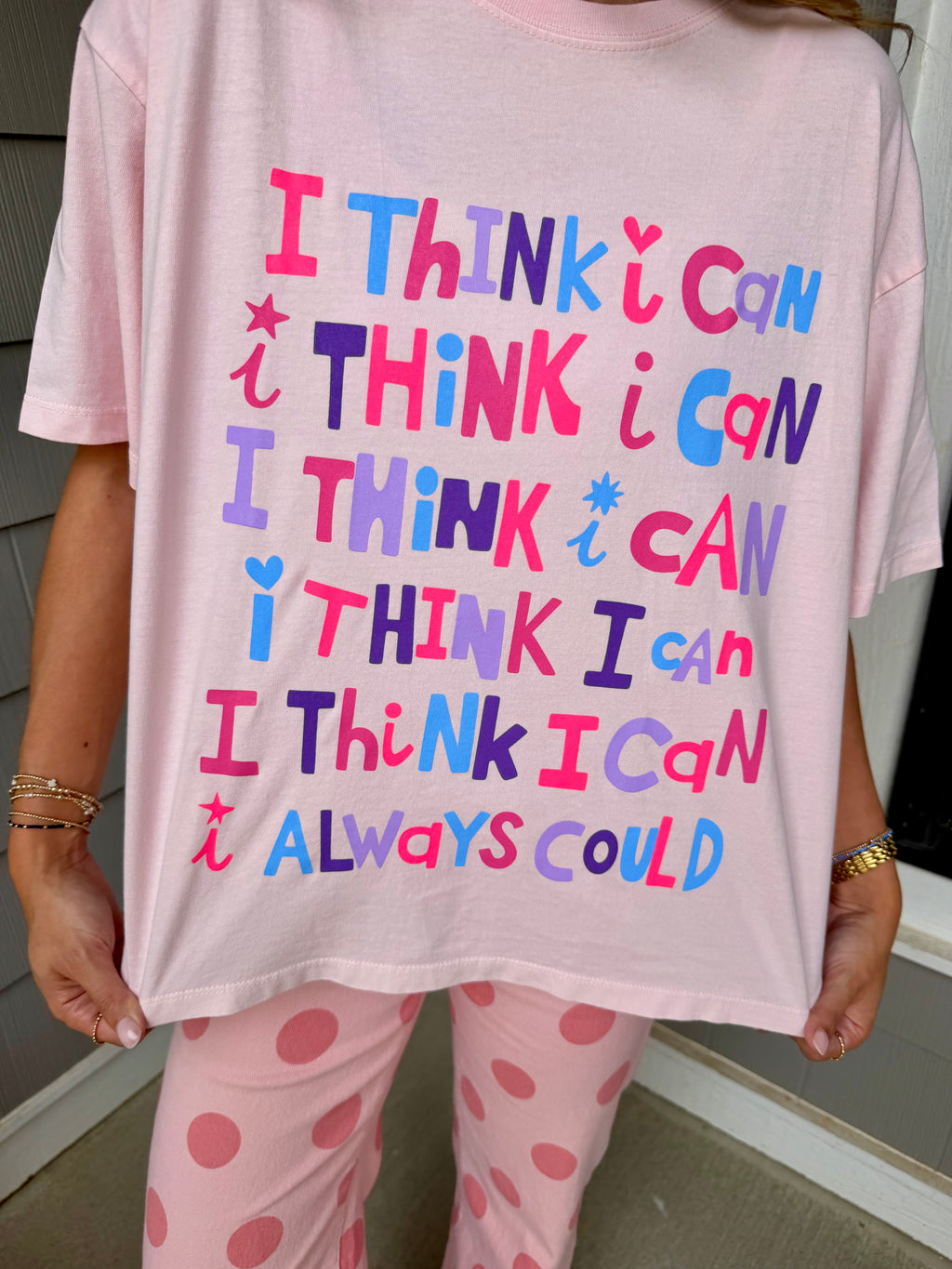 A person wearing a light pink oversized T-shirt with colorful text that reads 'I THINK I CAN I THINK I CAN I THINK I CAN I THINK I CAN I THINK I CAN I ALWAYS COULD' paired with pink polka-dot pajama pants.