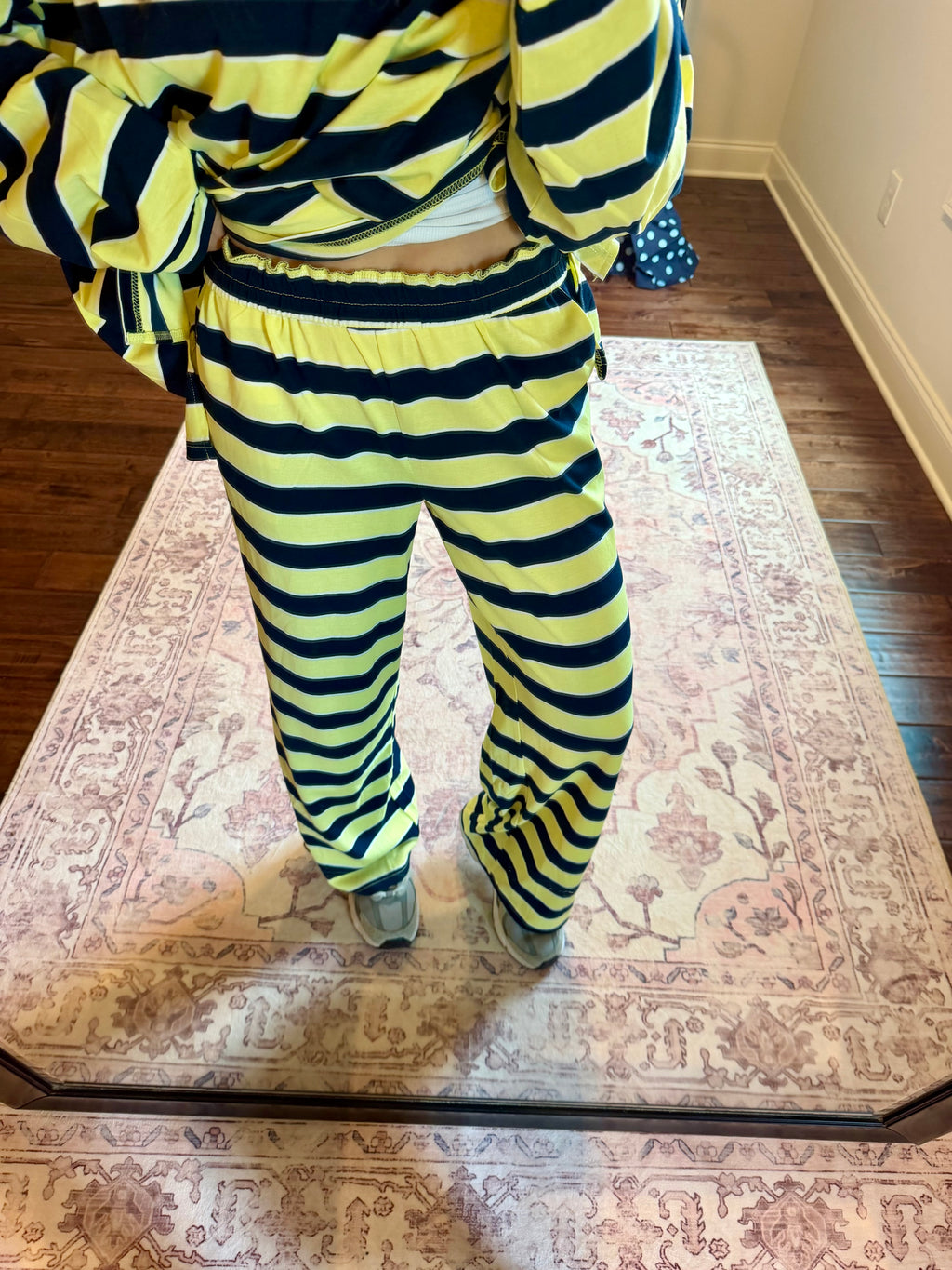 A person wearing a matching yellow and black striped lounge set, consisting of a cropped long-sleeve top and wide-leg pants, standing on a patterned area rug in a room with hardwood floors.