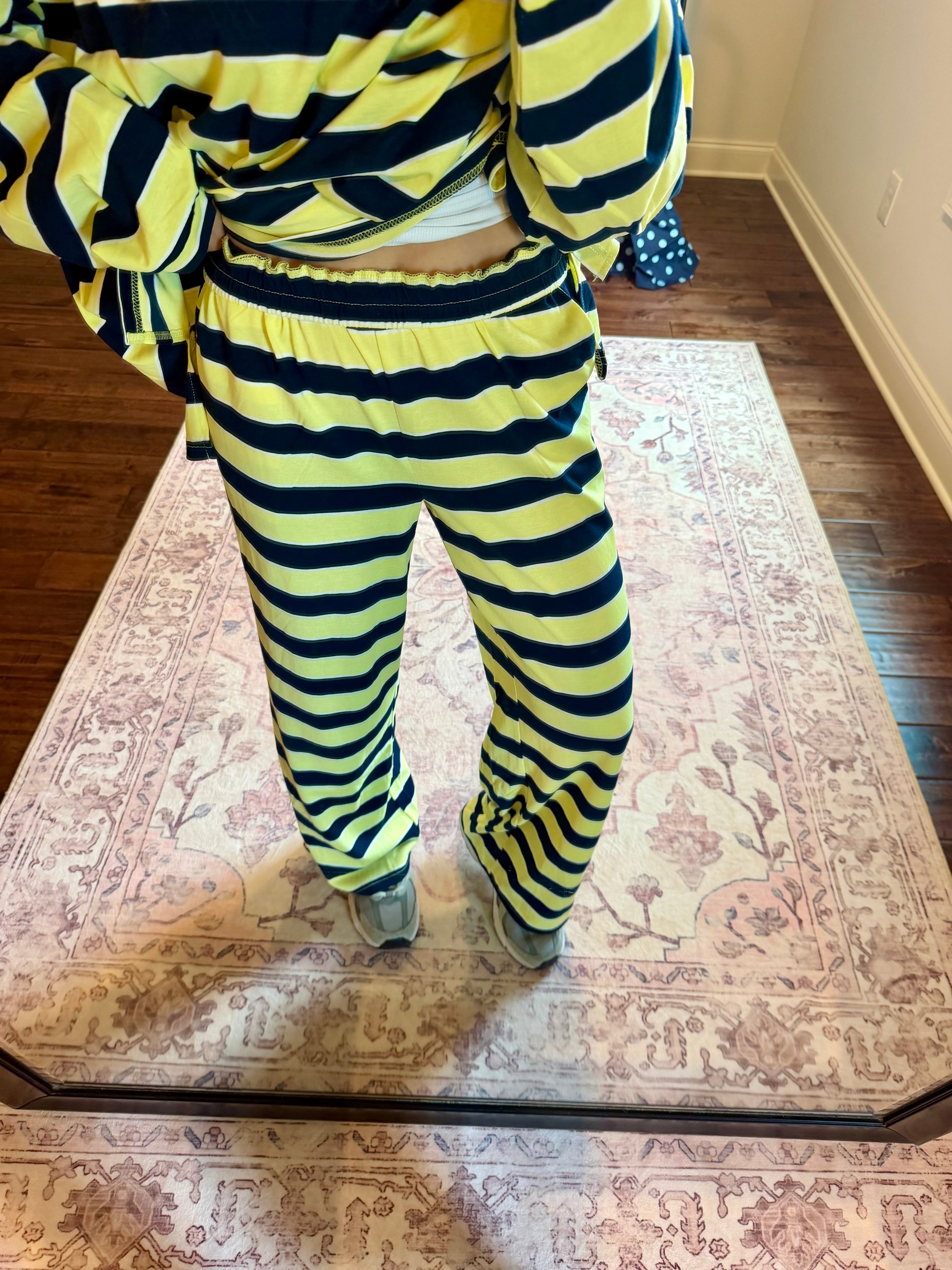 A person wearing a matching yellow and black striped lounge set, consisting of a cropped long-sleeve top and wide-leg pants, standing on a patterned area rug in a room with hardwood floors.