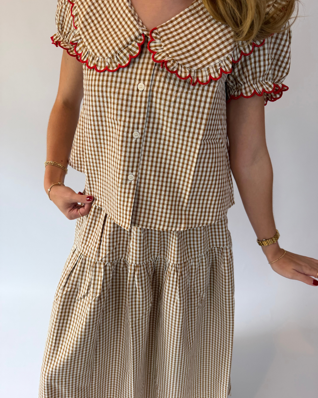 A woman wearing a brown and beige gingham two-piece set with a ruffled collar trimmed in red pom-pom, short puff sleeves, and a matching skirt.