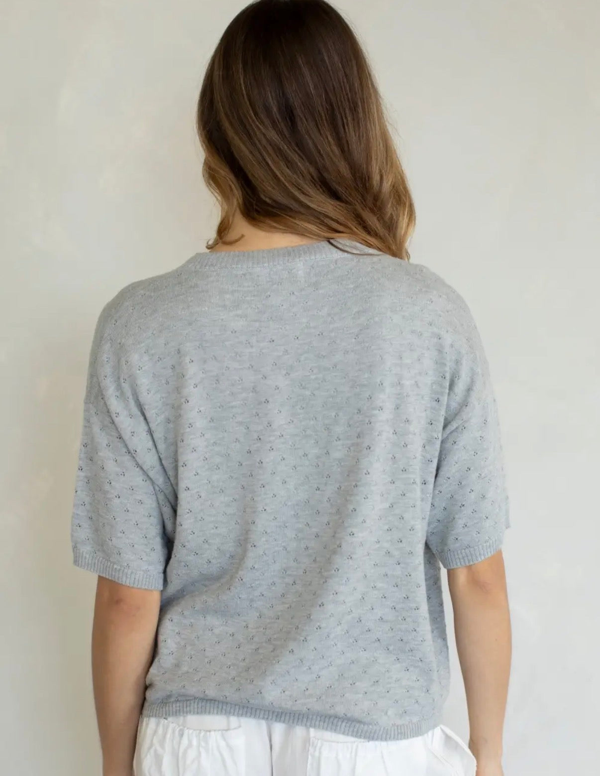 A woman viewed from behind wearing a light gray short-sleeve knit top with a subtle dotted texture and white pants, set against a plain off-white wall.