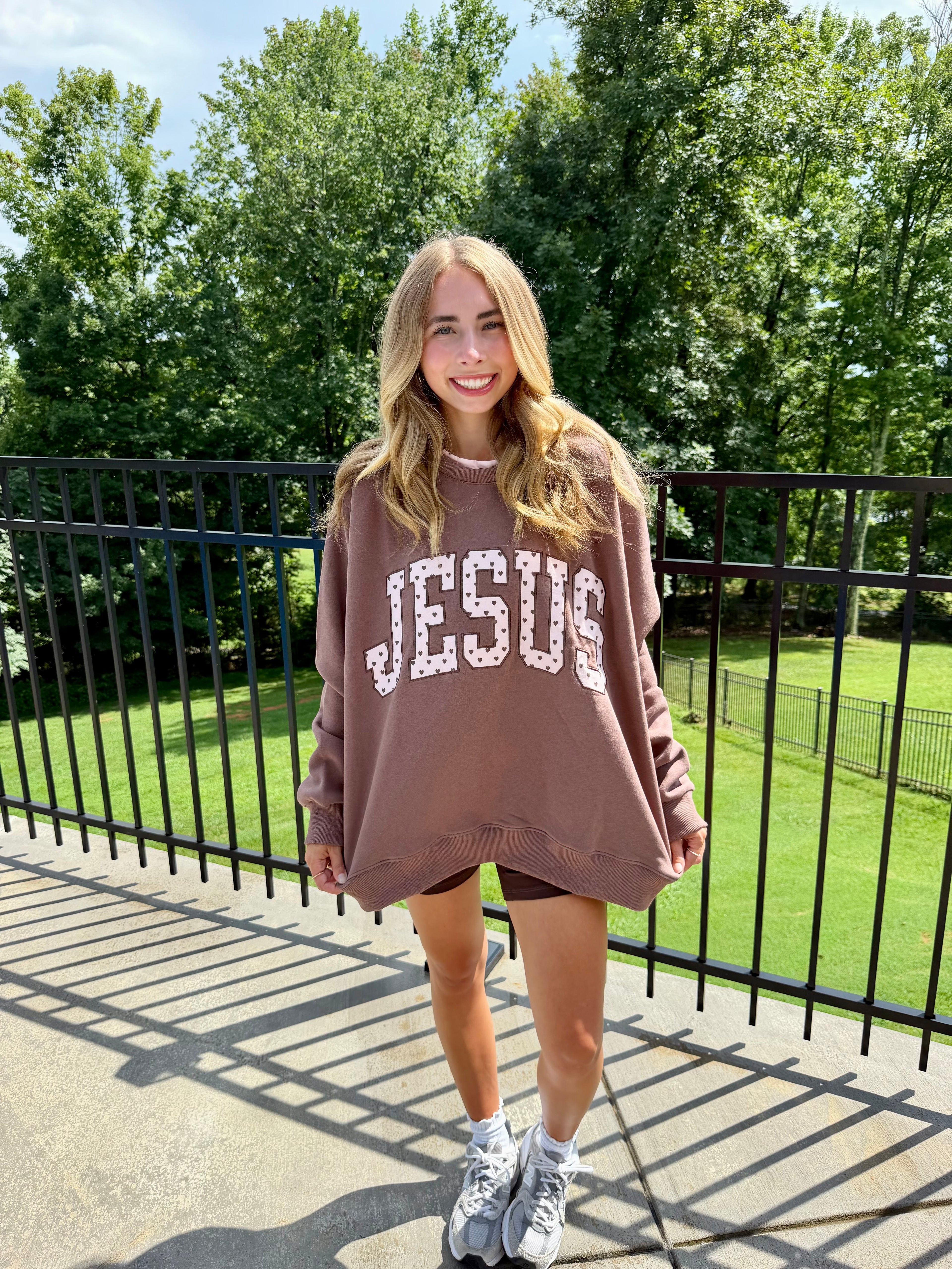 A woman stands outdoors on a concrete walkway wearing an oversized taupe sweatshirt with 'JESUS' printed across the front in white varsity-style letters outlined with small rhinestones. She is also wearing black shorts and white sneakers, with a black metal fence and green trees in the background.