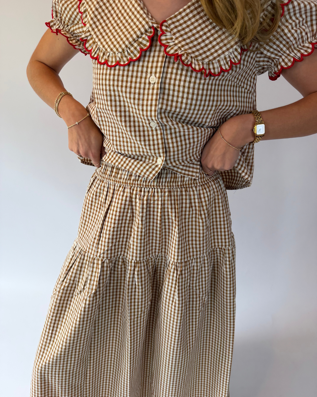 A person wearing a beige and brown gingham dress with red ruffle trim along the collar and sleeve edges, standing against a plain light background.