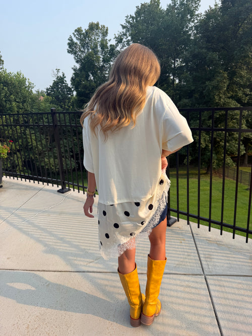 A woman viewed from behind wearing a cream-colored short-sleeve top, a white skirt with black polka dots, denim shorts, and bright yellow knee-high boots, standing on a concrete patio with a black metal railing and green trees in the background.