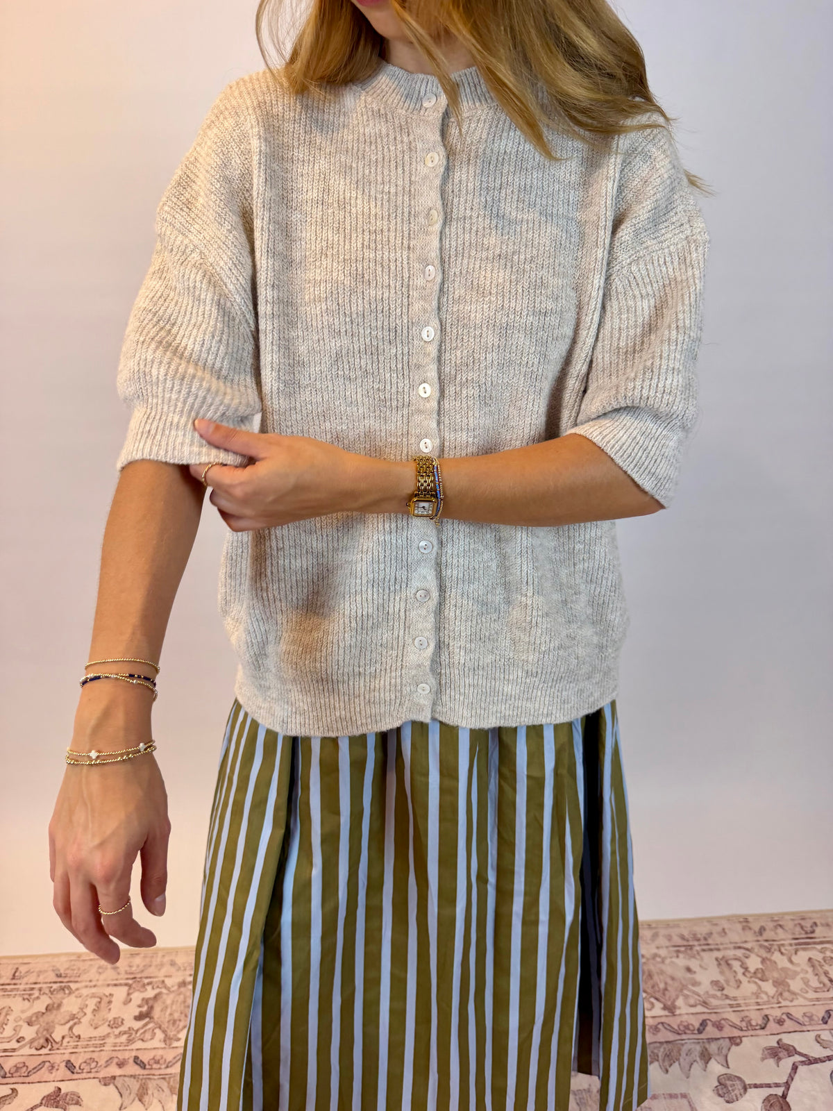 A woman wearing a cream-colored, short-sleeved ribbed knit button-up cardigan and a green and white vertically striped skirt, standing against a plain light background.