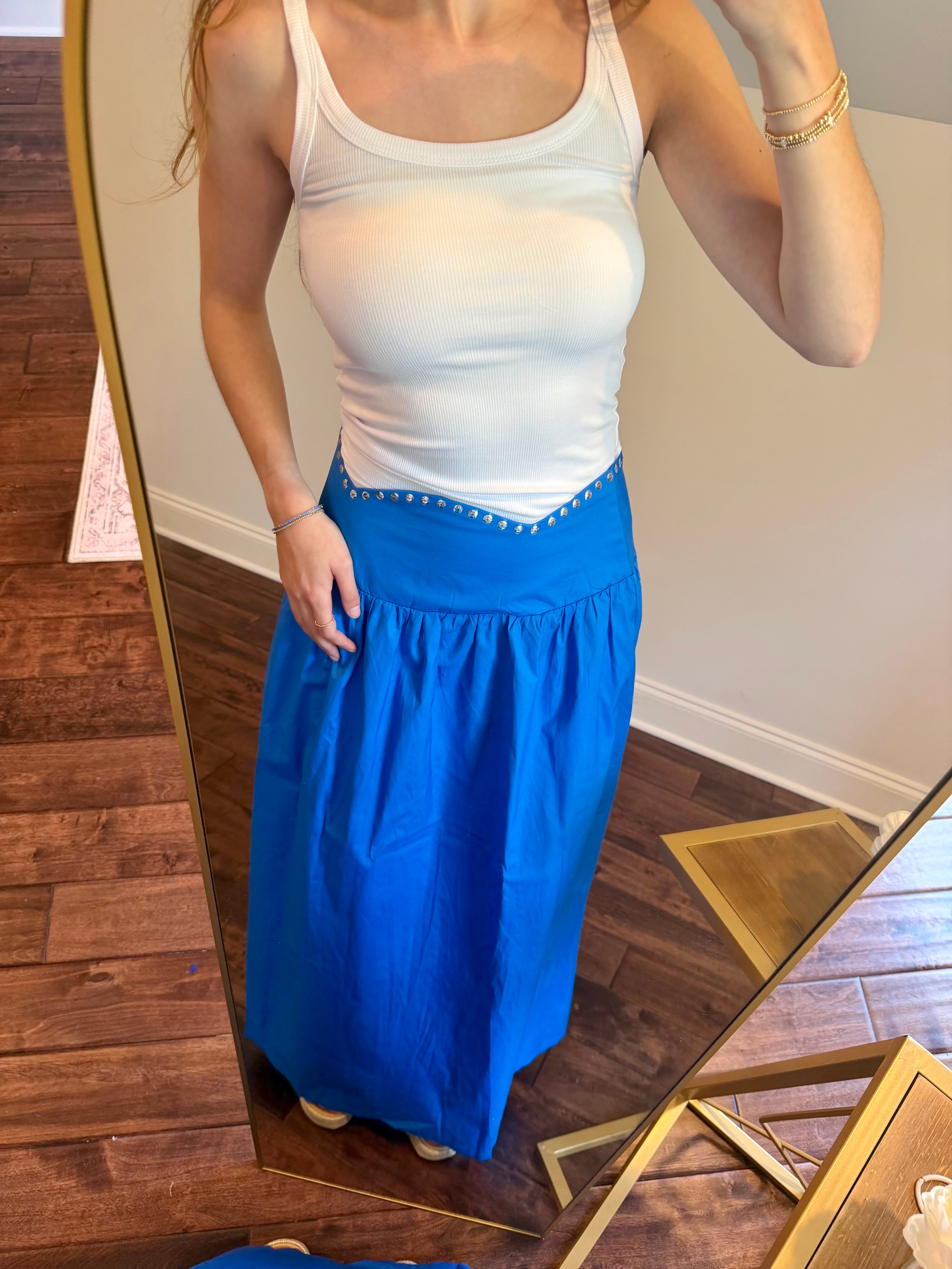 Person wearing a white tank top and blue skirt standing in front of a mirror.