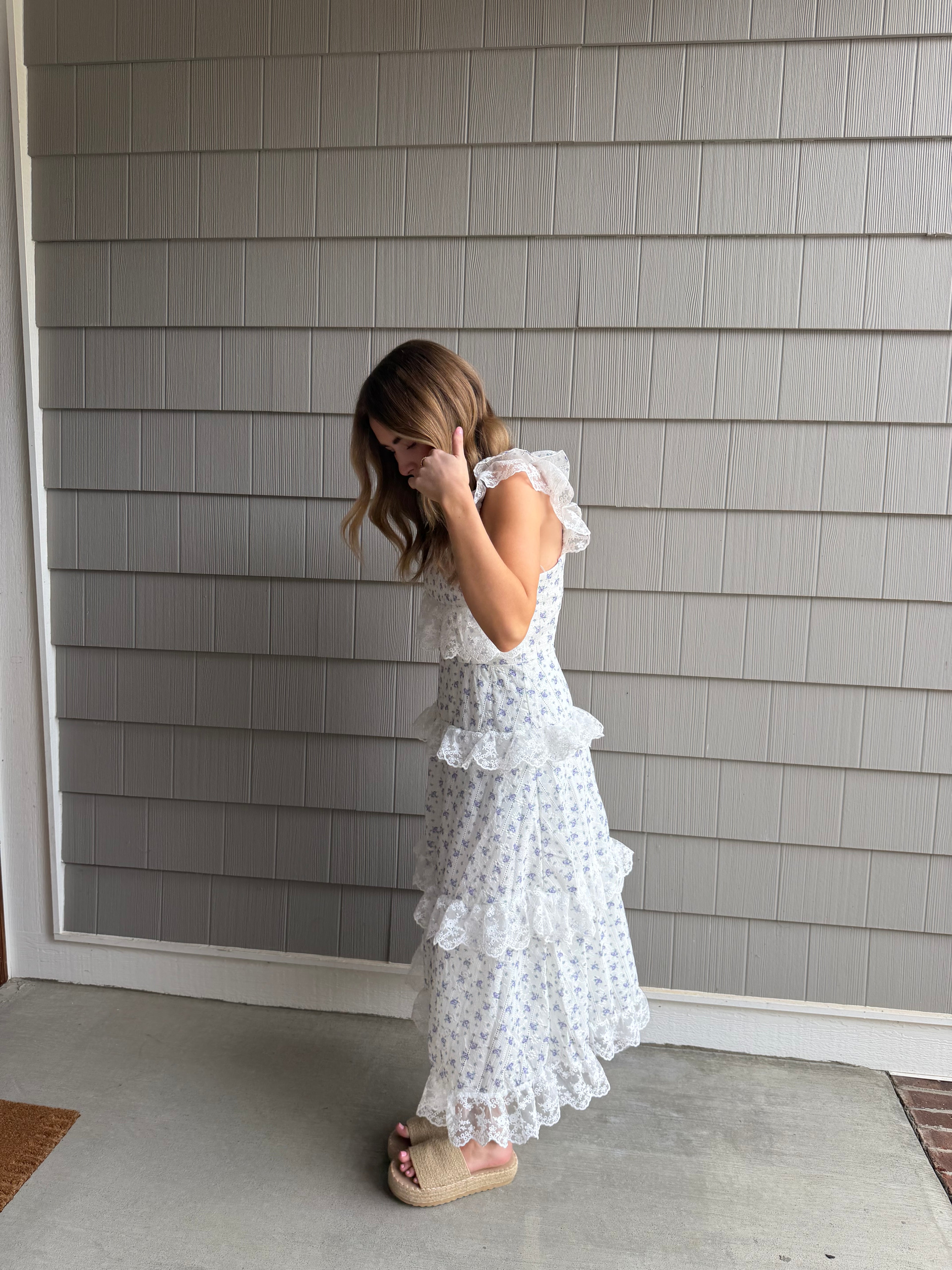 Woman wearing a white dress with ruffles standing against a gray paneled wall.