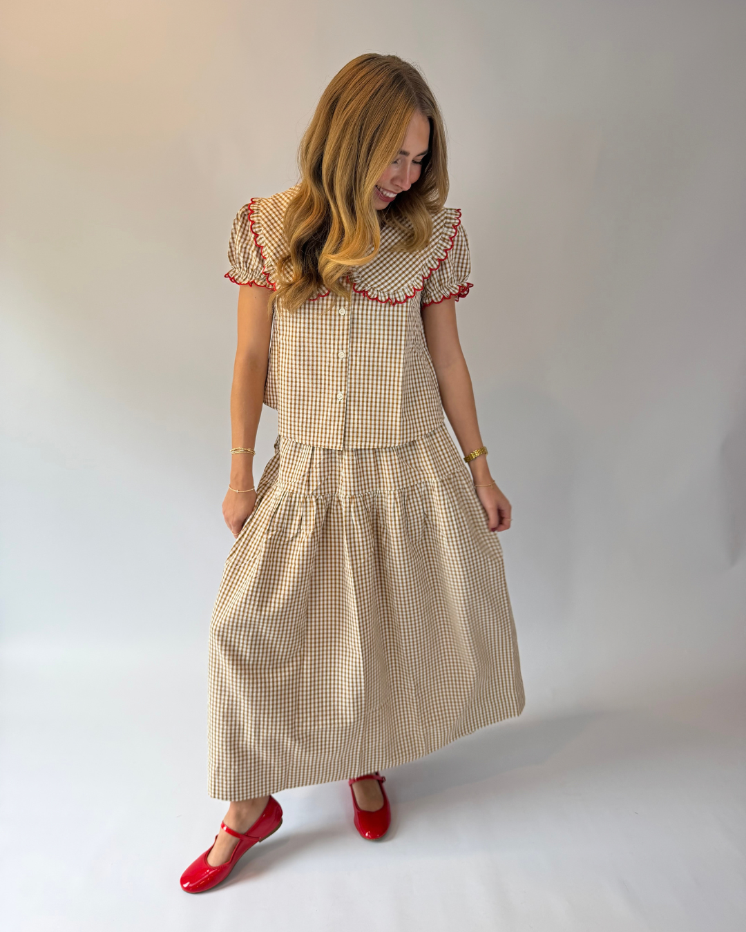 A woman modeling a beige and white gingham midi dress with red tassel trim on the collar and sleeves, paired with red Mary Jane shoes, against a plain light gray studio background.