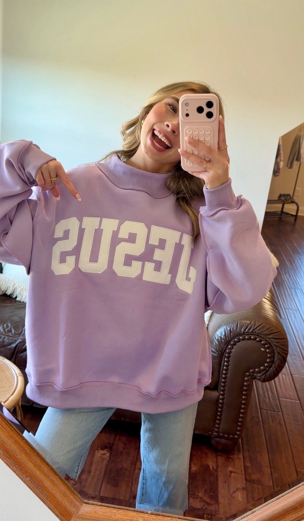 Person wearing a purple sweatshirt with 'Jesus' text, standing in a room with wooden flooring and a mirror.