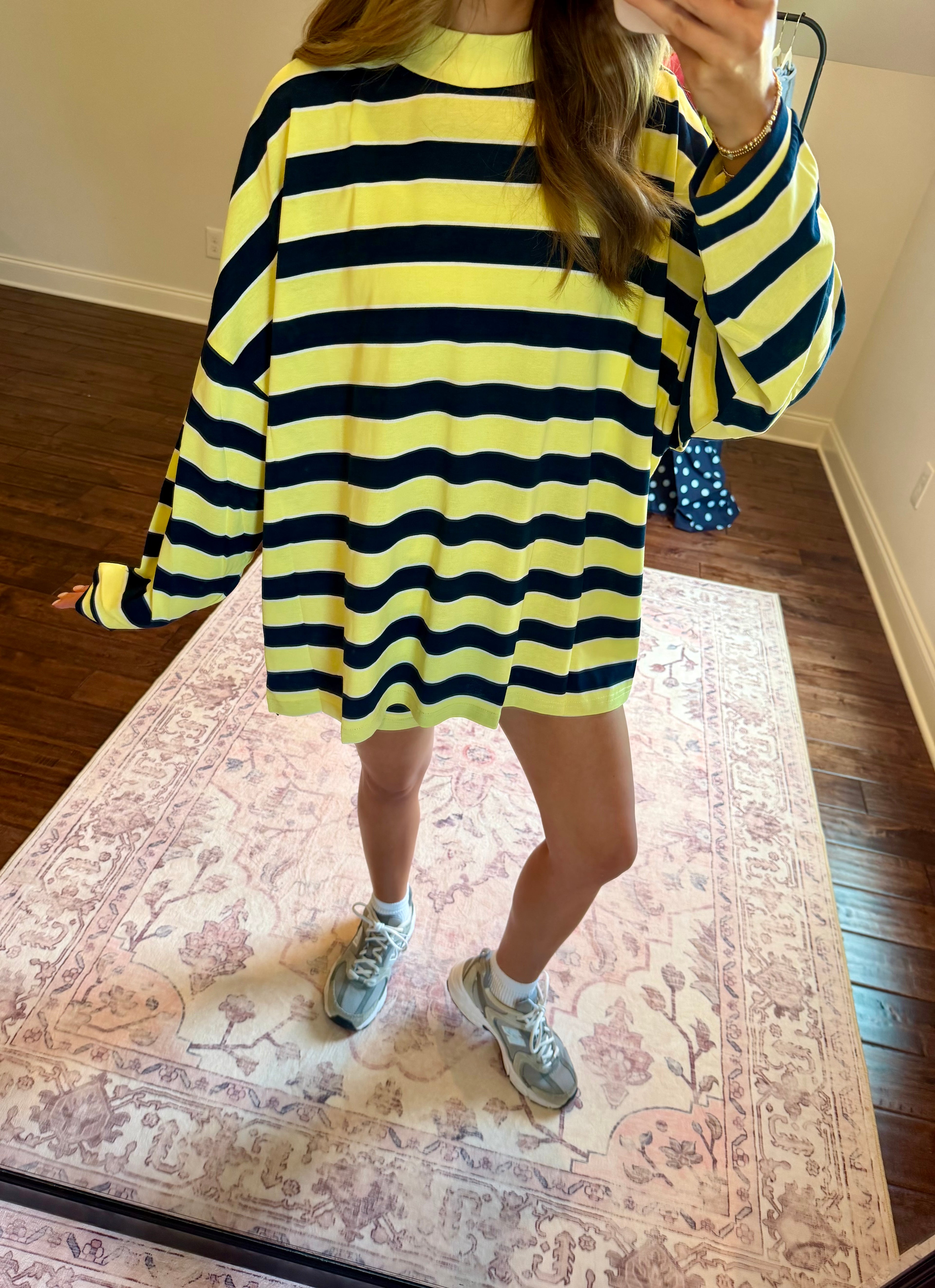 A person wearing an oversized long-sleeve knit top with bold horizontal neon yellow and black stripes, standing on a patterned rug in a room with hardwood floors.
