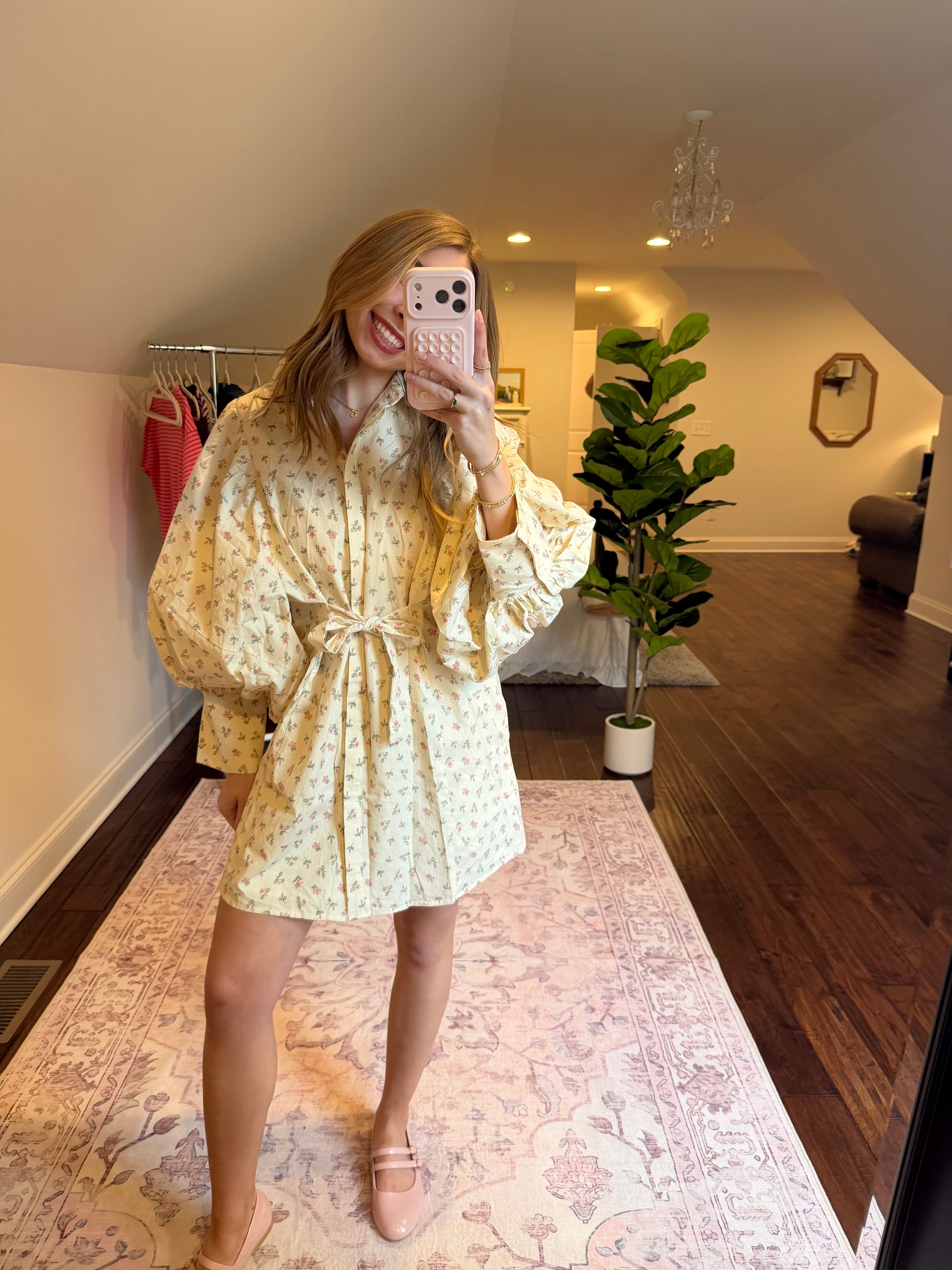 Woman in a floral dress standing on a patterned rug in a room with a plant and furniture.