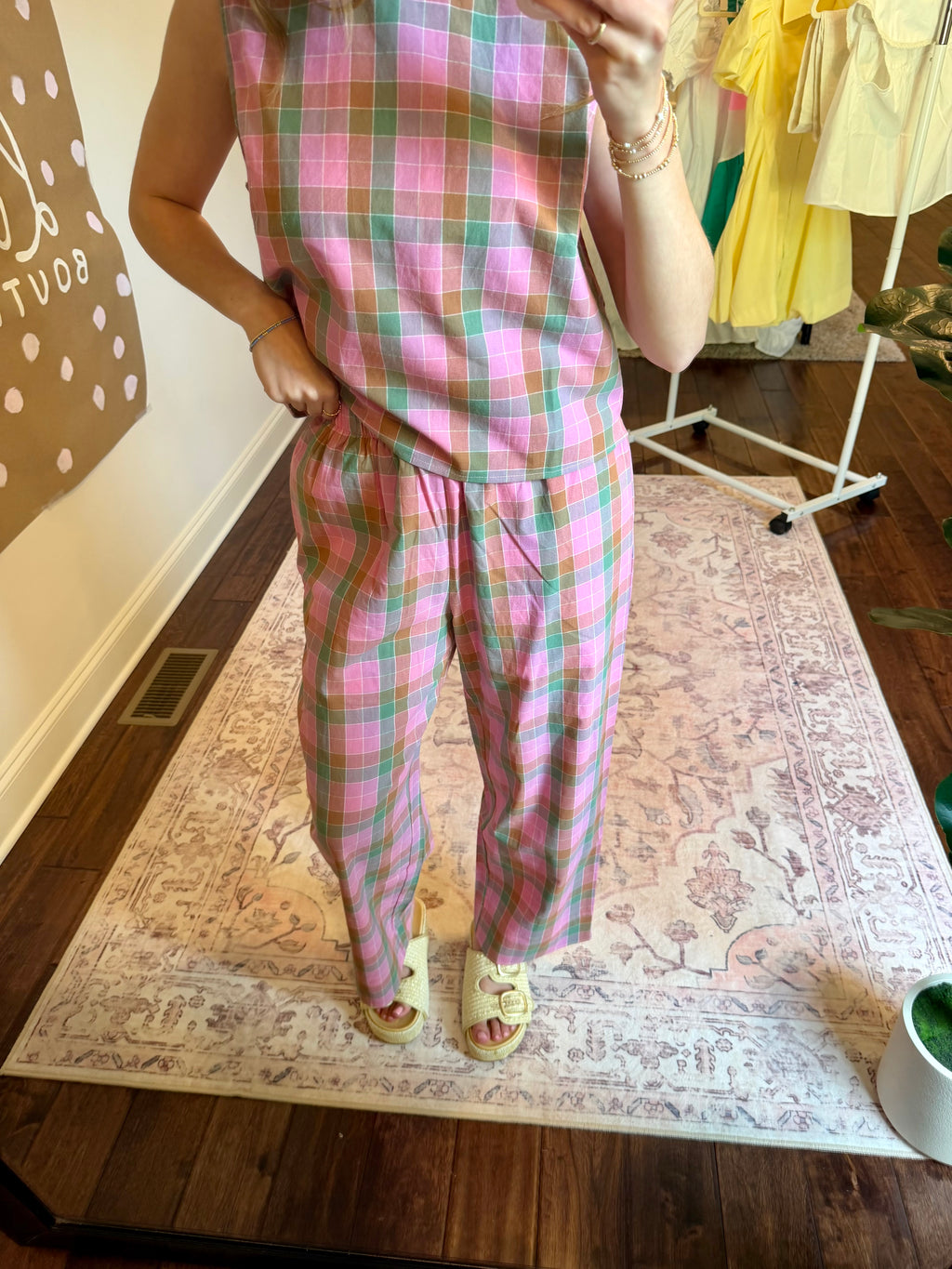 Person wearing a pink plaid outfit taking a mirror selfie in a room with a clothing rack in the background.
