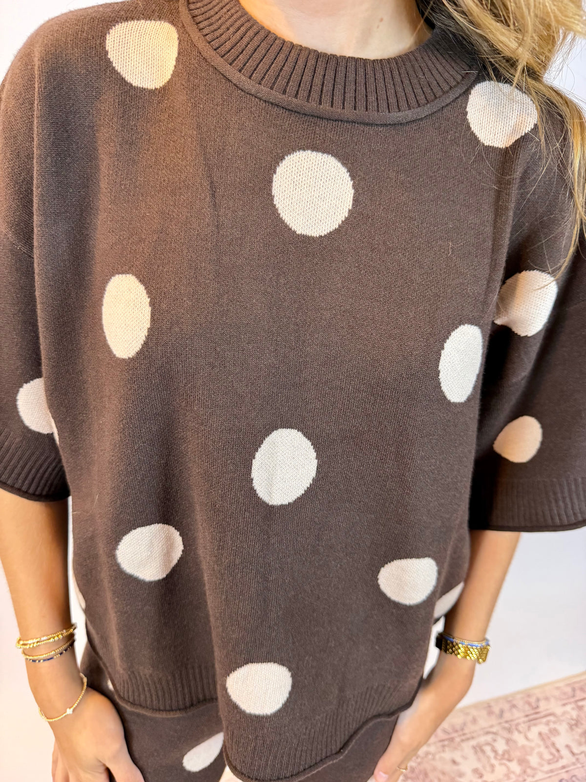A close-up of a person wearing an oversized brown knit sweater with large cream polka dots. The photo is cropped from the neck to the hips, showing the ribbed crew neck, short sleeves, and patterned fabric against a light indoor background.
