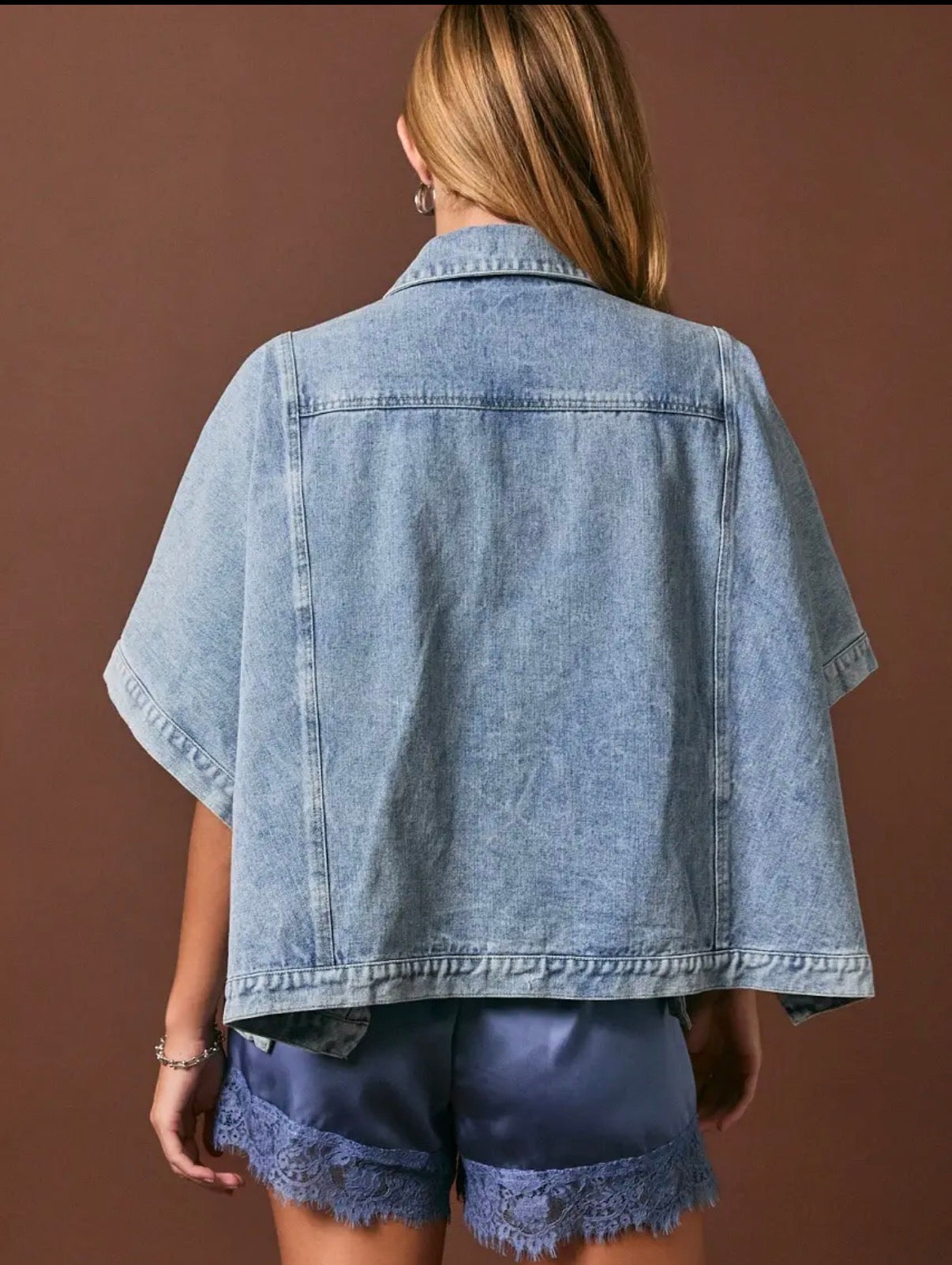 A woman shown from the back wearing a light-wash blue denim short-sleeve cropped jacket and dark denim shorts with frayed hems, set against a solid brown studio background.