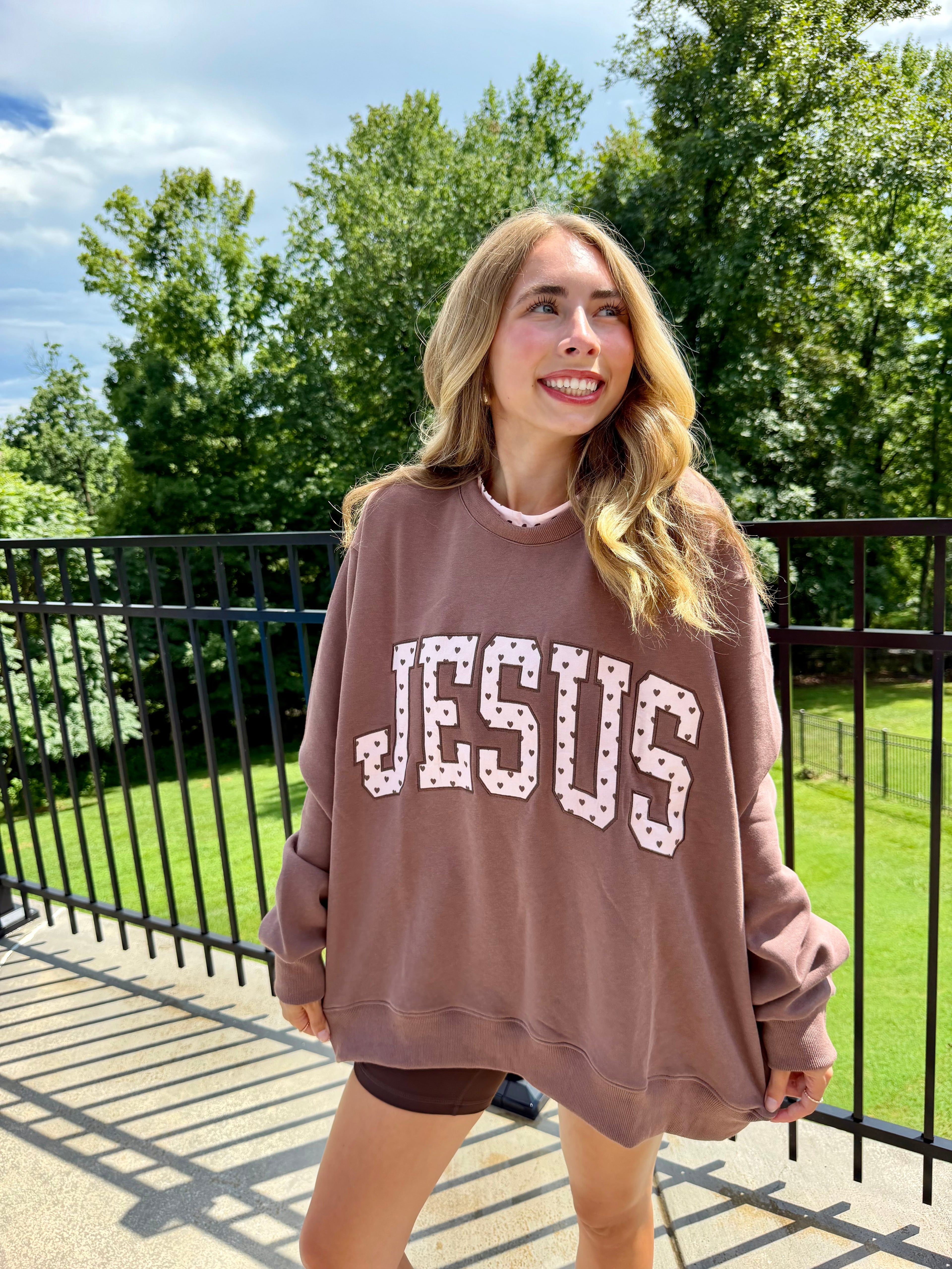 A woman wearing a brownish-pink oversized sweatshirt with 'JESUS' printed across the front in a patterned font, standing outdoors on a balcony with trees and grass in the background.