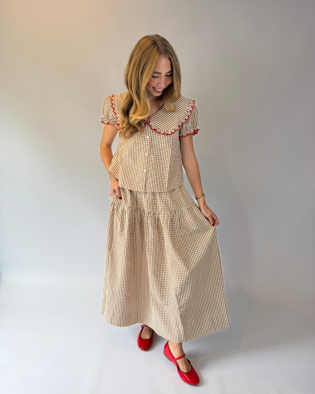 A woman modeling a vintage-style two-piece dress set featuring a beige and white gingham pattern with red embroidered trim on the collar and sleeve cuffs, paired with red Mary Jane shoes, against a neutral studio backdrop.