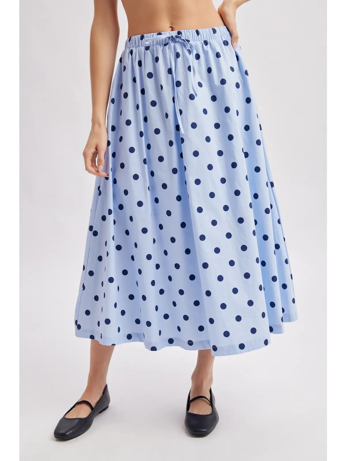 A model wearing a light blue midi skirt with a dark blue polka dot pattern, paired with black flat shoes, shown against a plain light gray background.