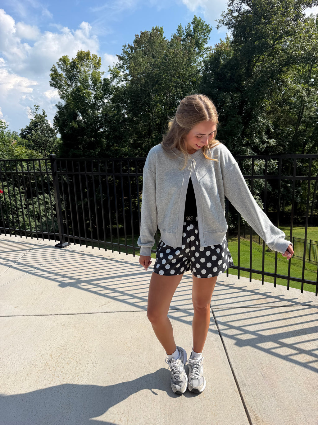 A woman stands outdoors on a concrete walkway wearing a light gray zip-up hoodie, a black top, black shorts with white polka dots, and white sneakers. Trees, grass, and a black metal fence are visible in the background under a blue sky.