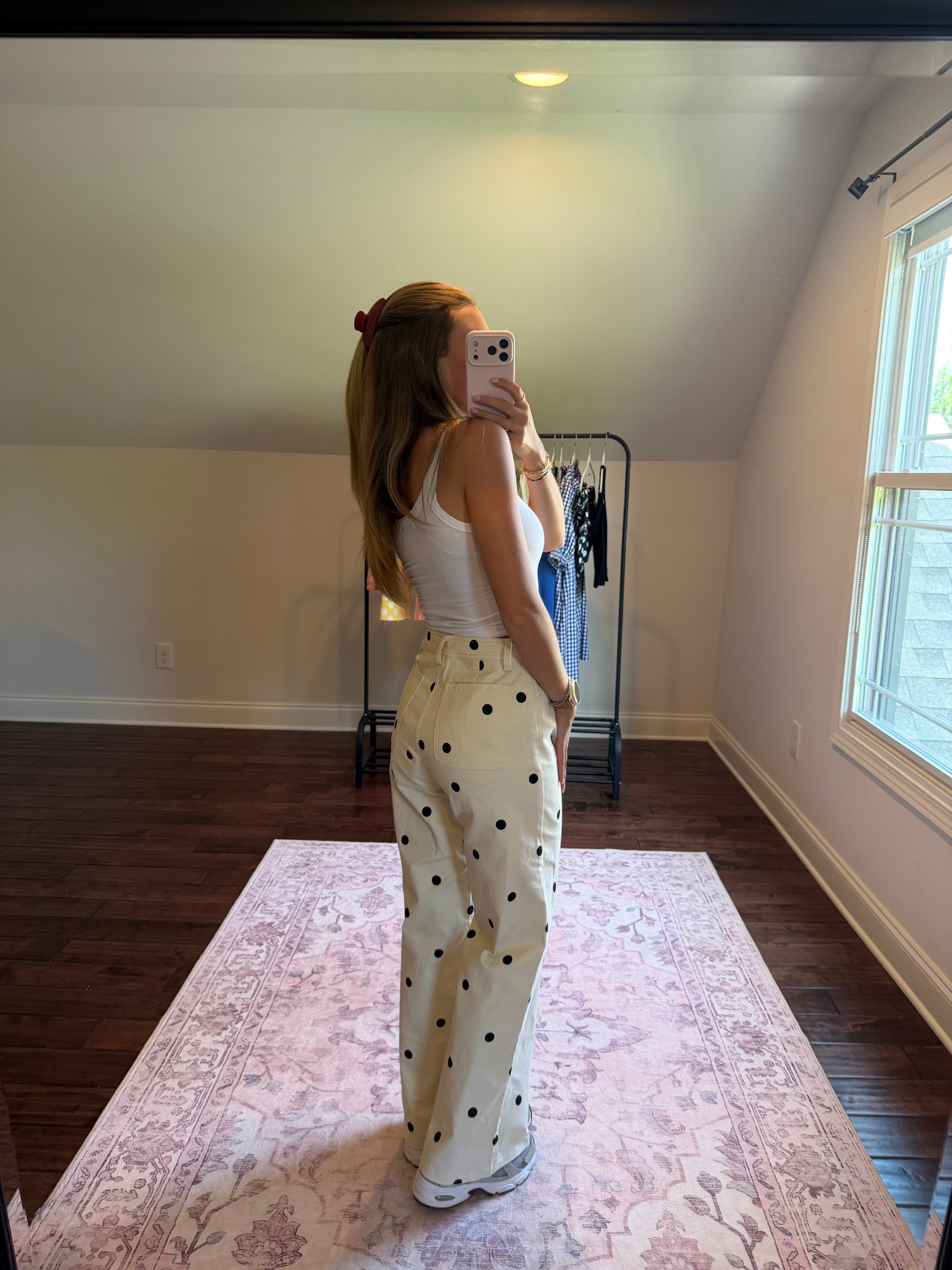 A woman takes a mirror selfie in a room with a slanted ceiling, wearing a white tank top and cream wide-leg pants with black polka dots. She is standing on a pink patterned rug with dark wood floors and a clothing rack in the background.