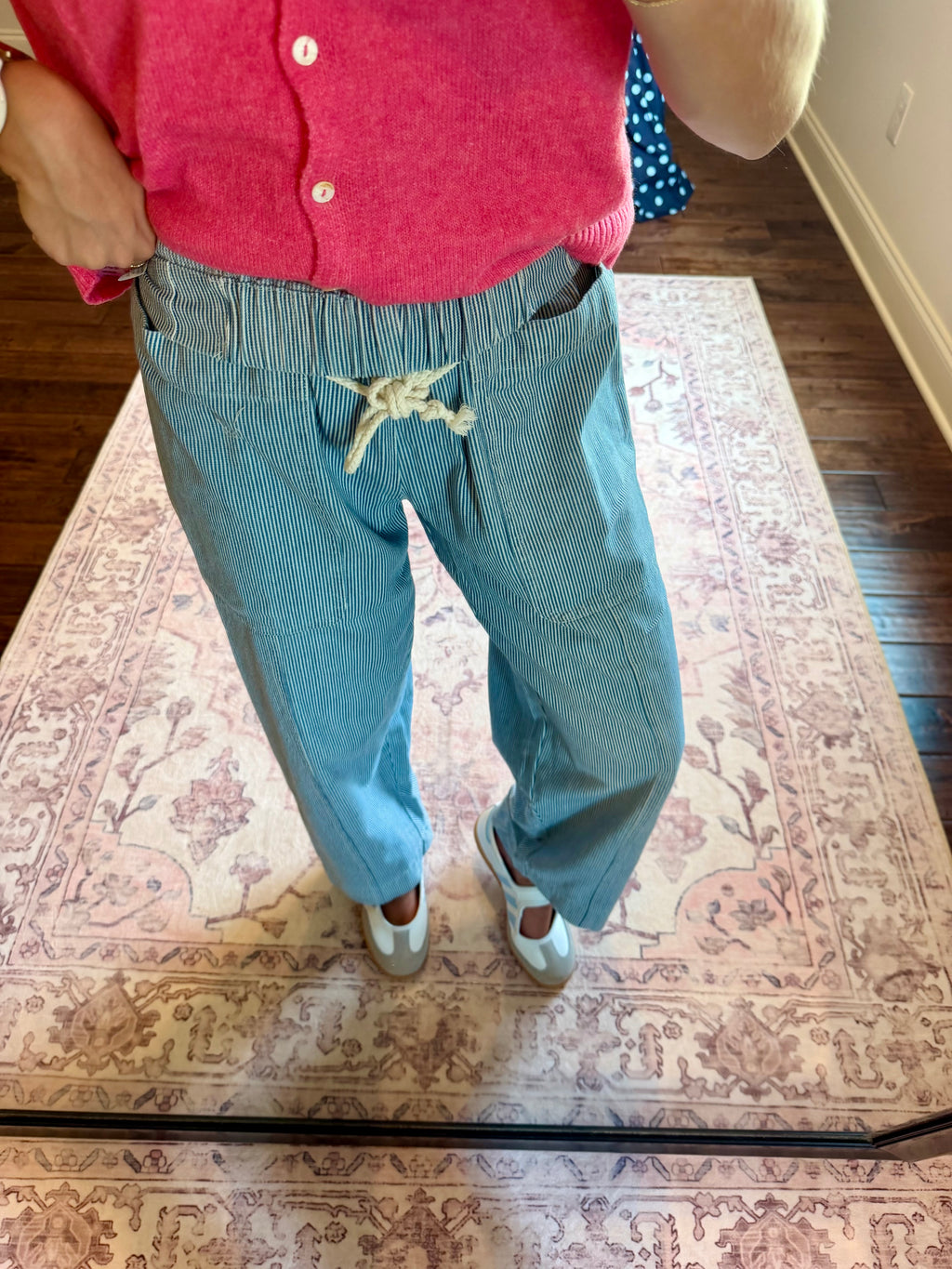 A person wearing light blue corduroy pants with a white drawstring, paired with a pink cardigan and white sneakers, standing on a patterned rug in a home setting.