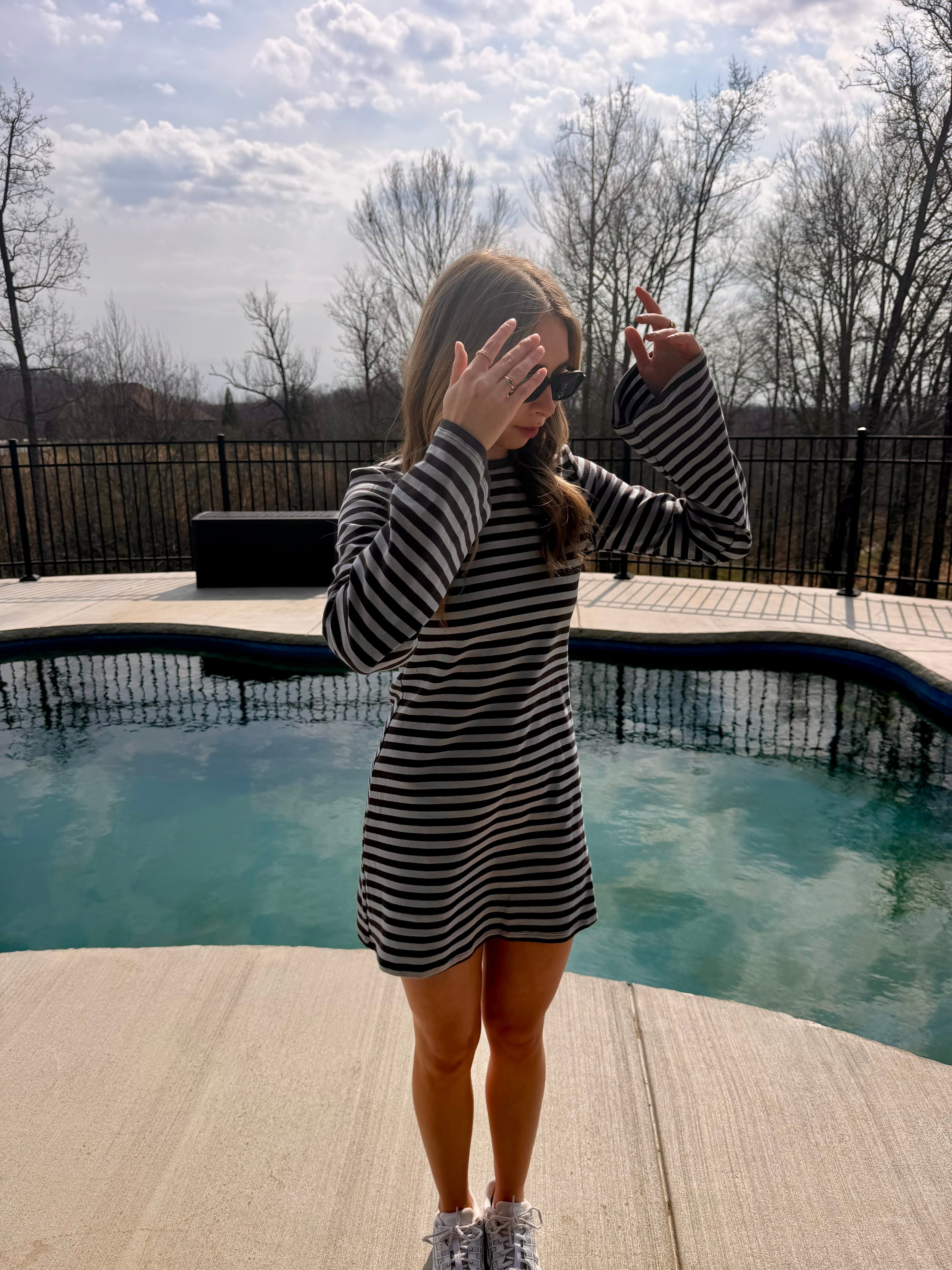 Woman in a striped dress standing by a pool with trees and a cloudy sky in the background