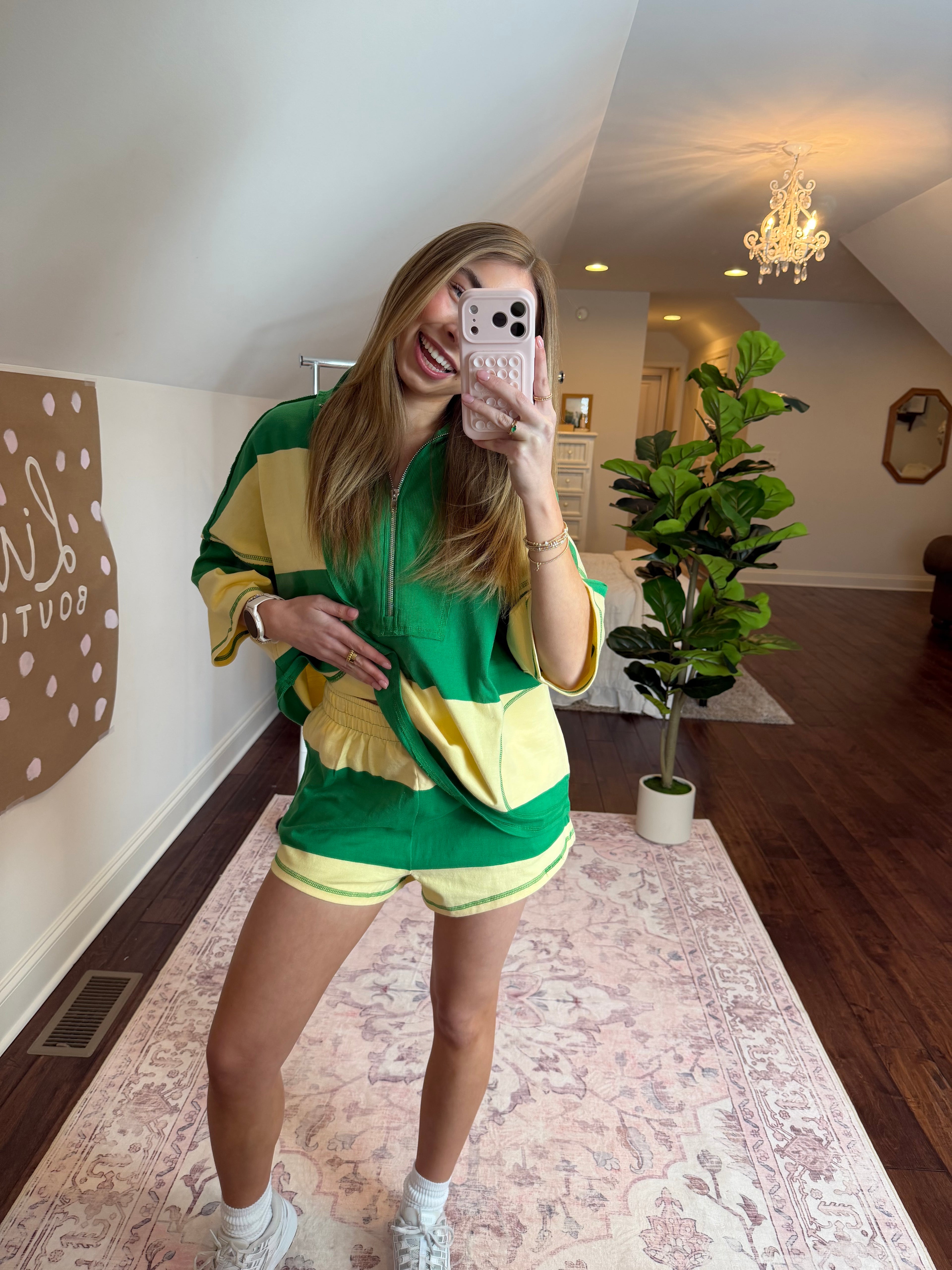 Person wearing a green and yellow outfit taking a selfie in a room with plants and a patterned rug.