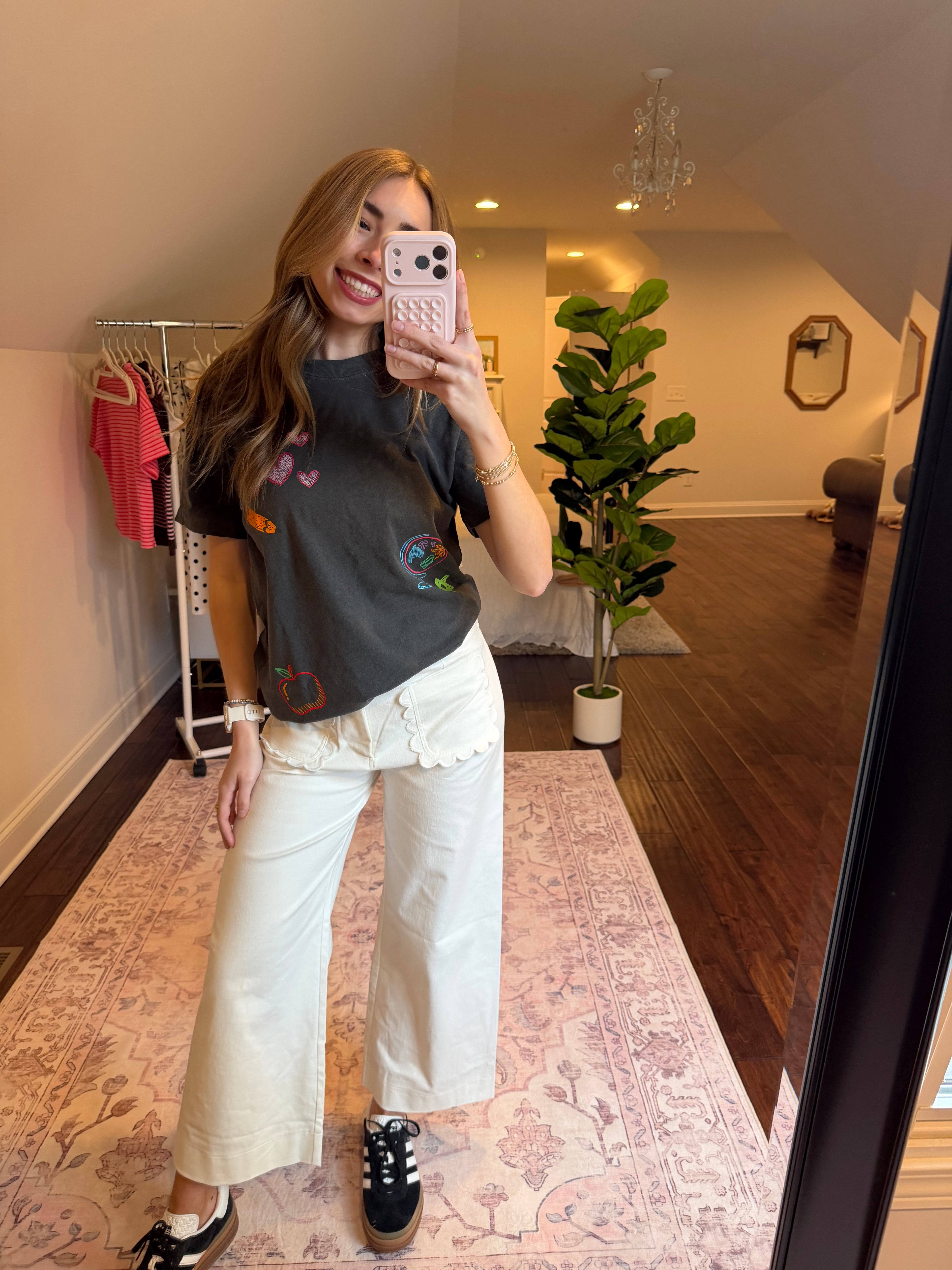 Person taking a mirror selfie wearing a dark t-shirt with colorful designs and light-colored pants in a room.