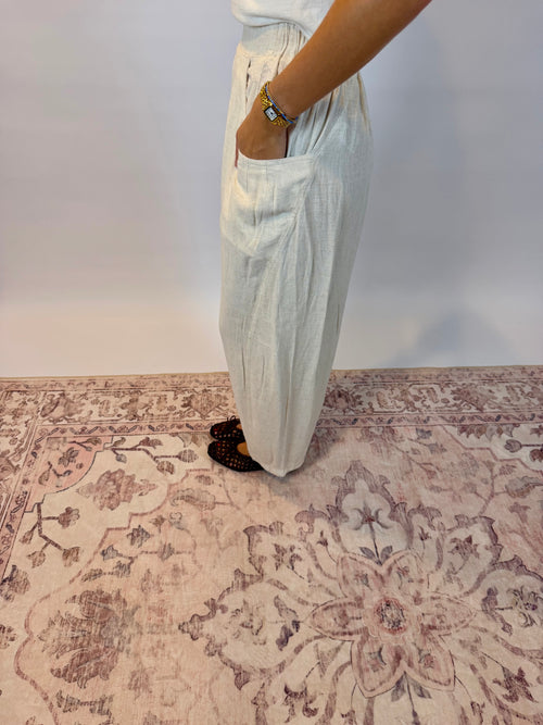 A person wearing light beige wide-leg linen pants with side pockets, standing on a distressed beige and mauve patterned area rug against a plain white wall.