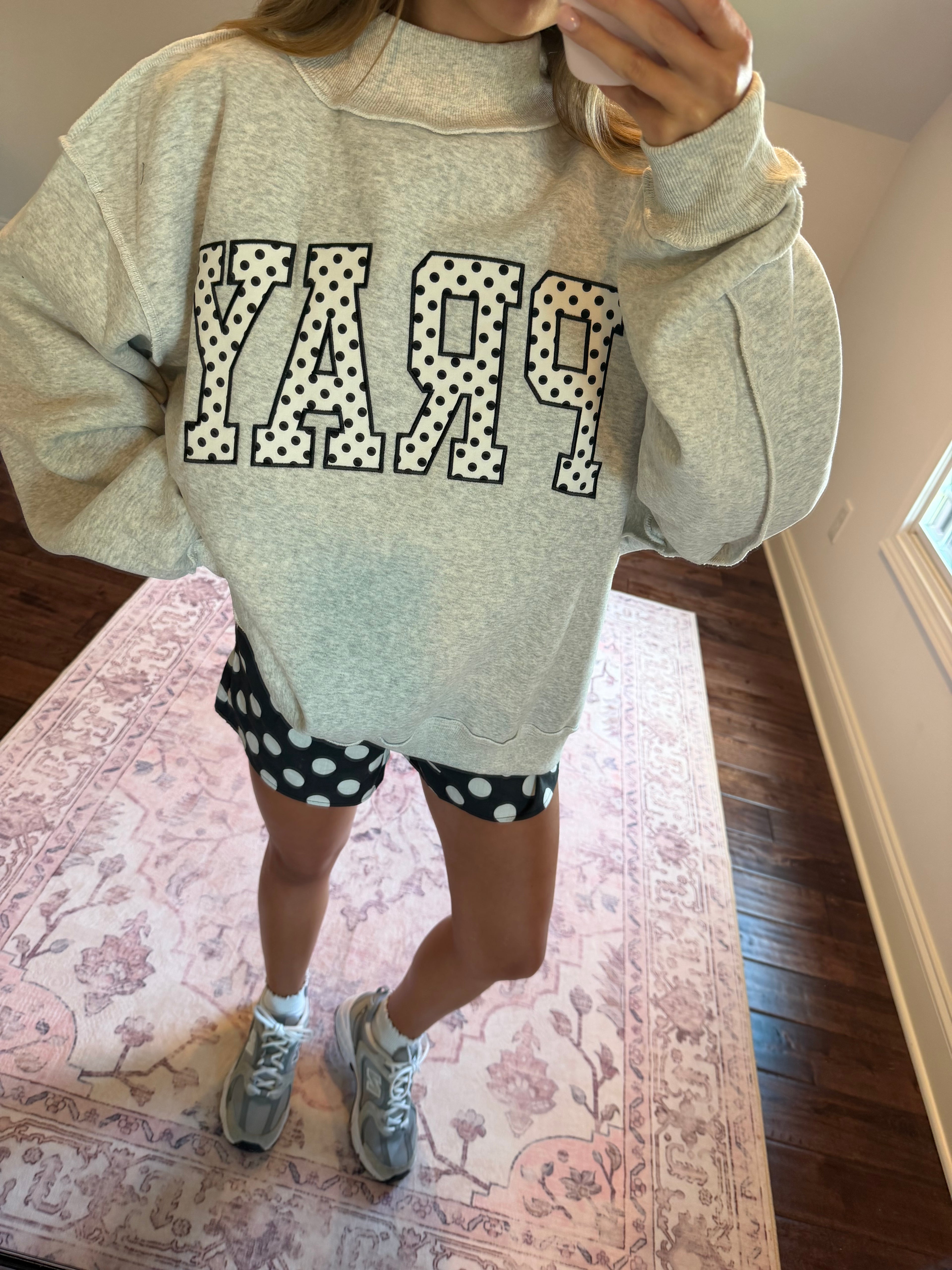A person wearing an oversized light heather gray sweatshirt with large polka-dot outlined letters spelling 'PRAY' across the front, paired with black-and-white polka-dot shorts and gray sneakers, standing indoors on a patterned rug.