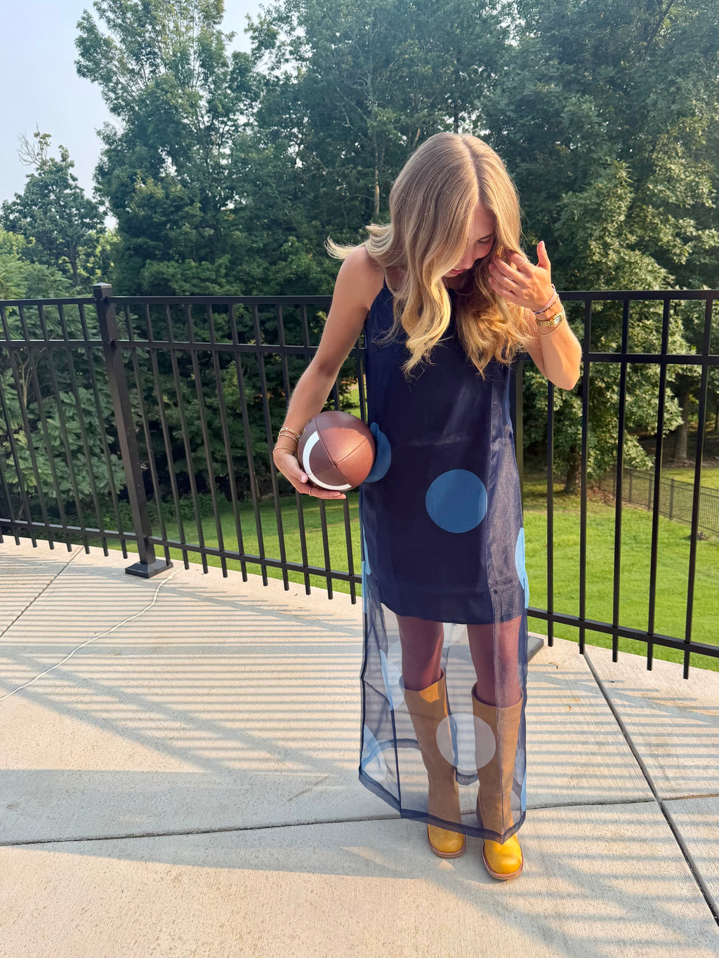 A woman stands on a concrete patio wearing a navy blue sleeveless dress with a sheer mesh overlay and blue circular graphic details, paired with brown boots. She holds a football in one hand, with a black metal fence and green trees in the background.