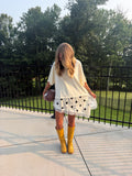 A woman stands on an outdoor patio wearing a cream oversized t-shirt, a white polka-dot mini skirt with lace trim, and yellow rain boots, holding a football. Trees and a black metal fence are visible in the background.