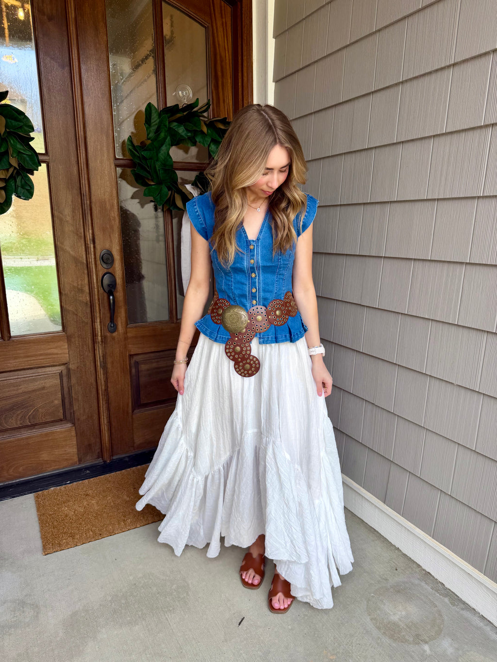 Woman in a blue top and white skirt standing outside a house.