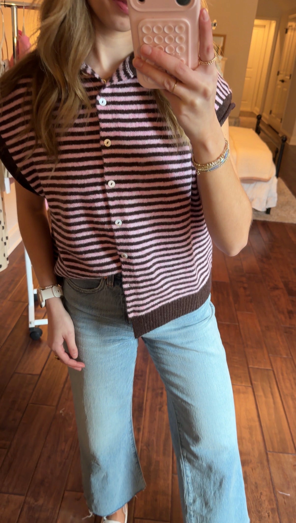 Person taking a mirror selfie wearing a pink and brown striped sweater vest and jeans in a bedroom.