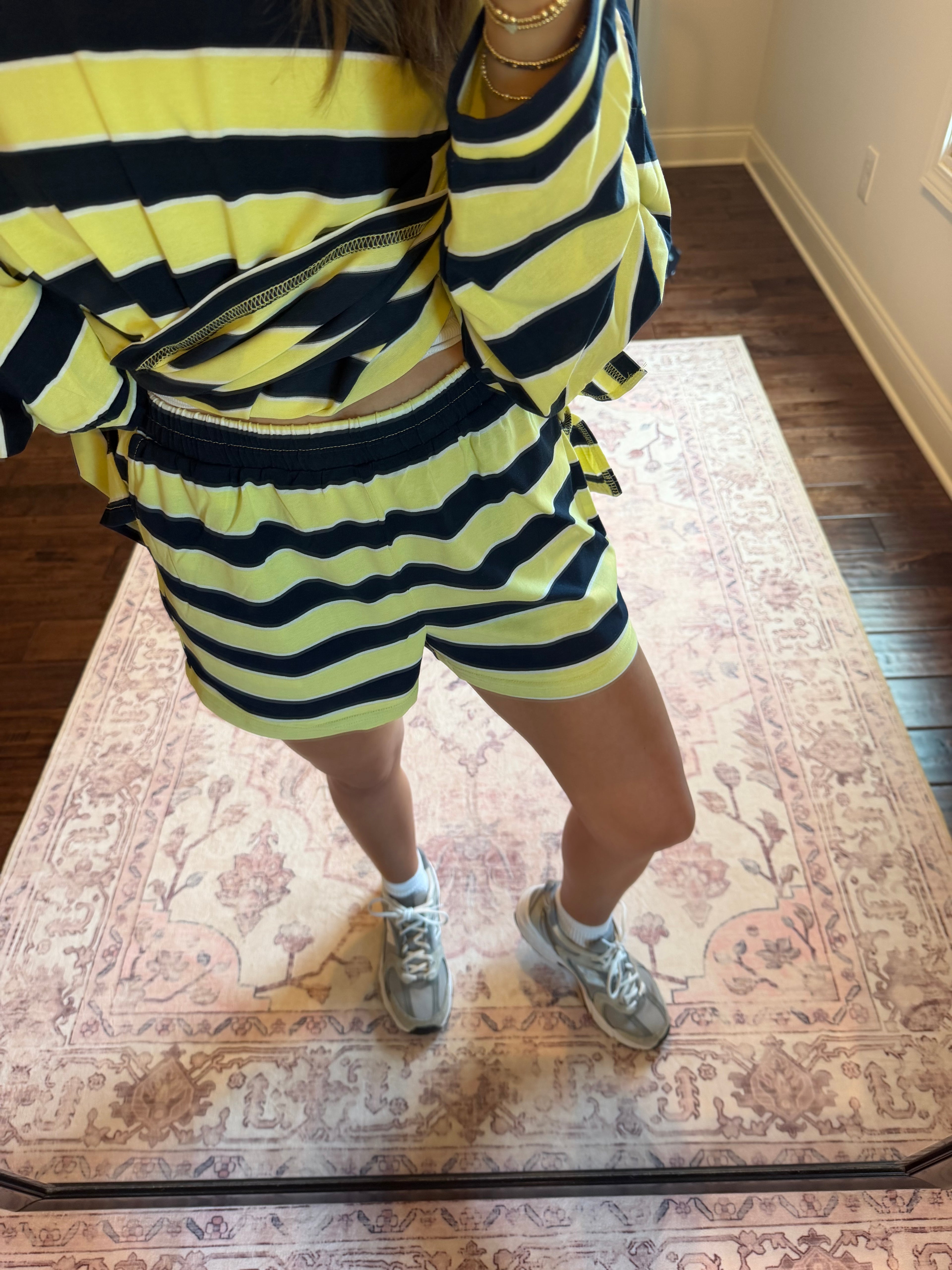A woman takes a mirror selfie in a room wearing a yellow and black striped oversized sweater and matching shorts. She is standing on a pink patterned area rug with hardwood floors, a window, and a clothing rack in the background.