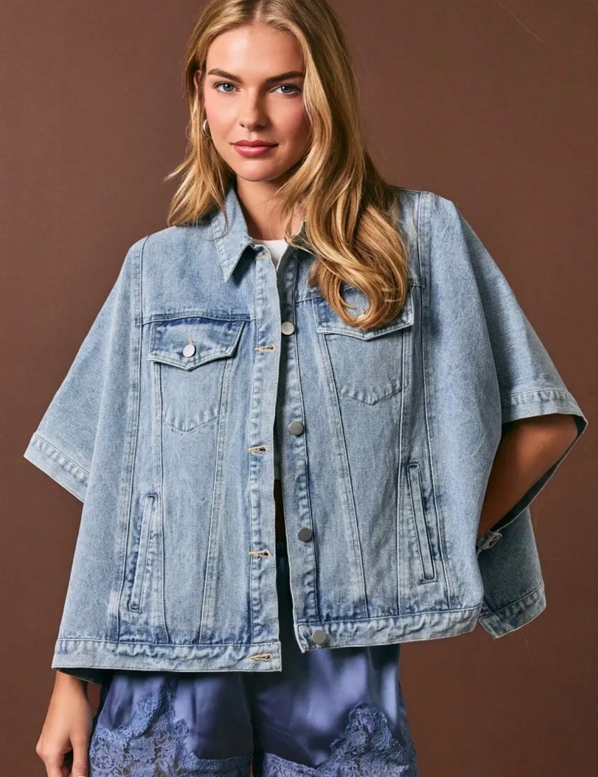 A woman wearing a light-wash denim short-sleeve button-up jacket with chest flap pockets, photographed against a solid brown studio background.