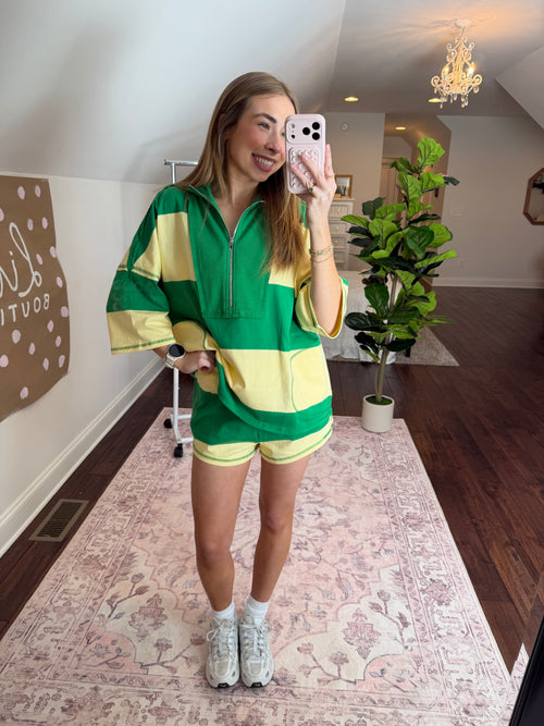 Person wearing a green and yellow outfit taking a selfie in a room with plants and a patterned rug.