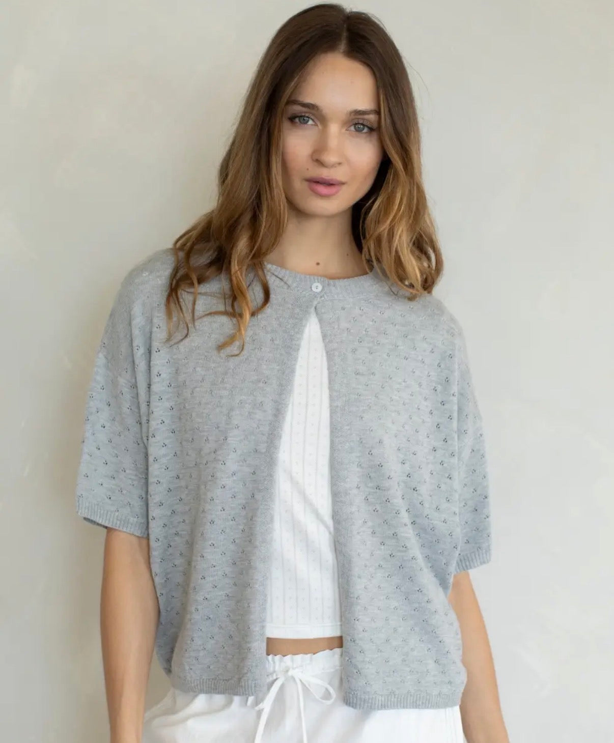 A woman wearing a light gray short-sleeve knit cardigan with tiny speckled details over a white ribbed crop top and white drawstring pants, standing against a plain light gray wall.
