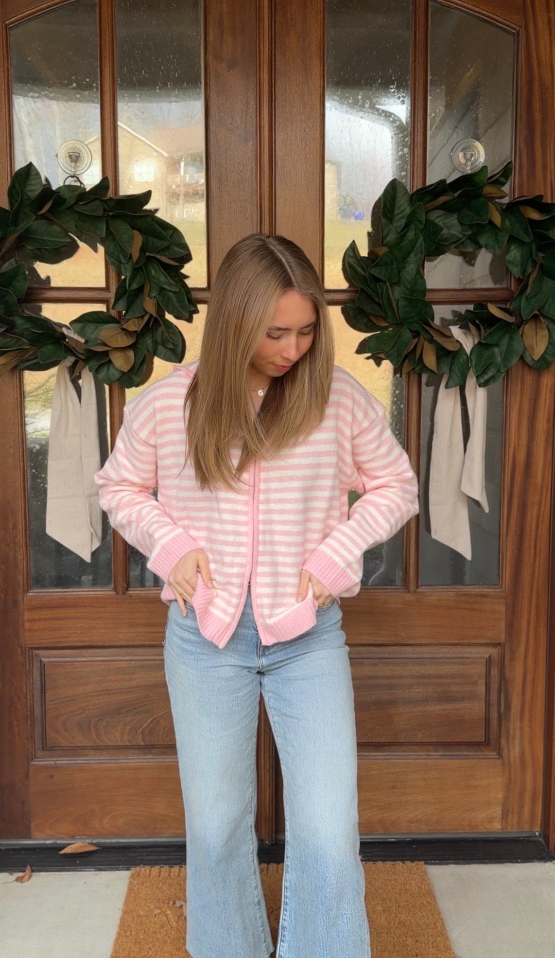Person wearing a pink striped sweater and light blue jeans standing in front of a wooden door with wreaths.
