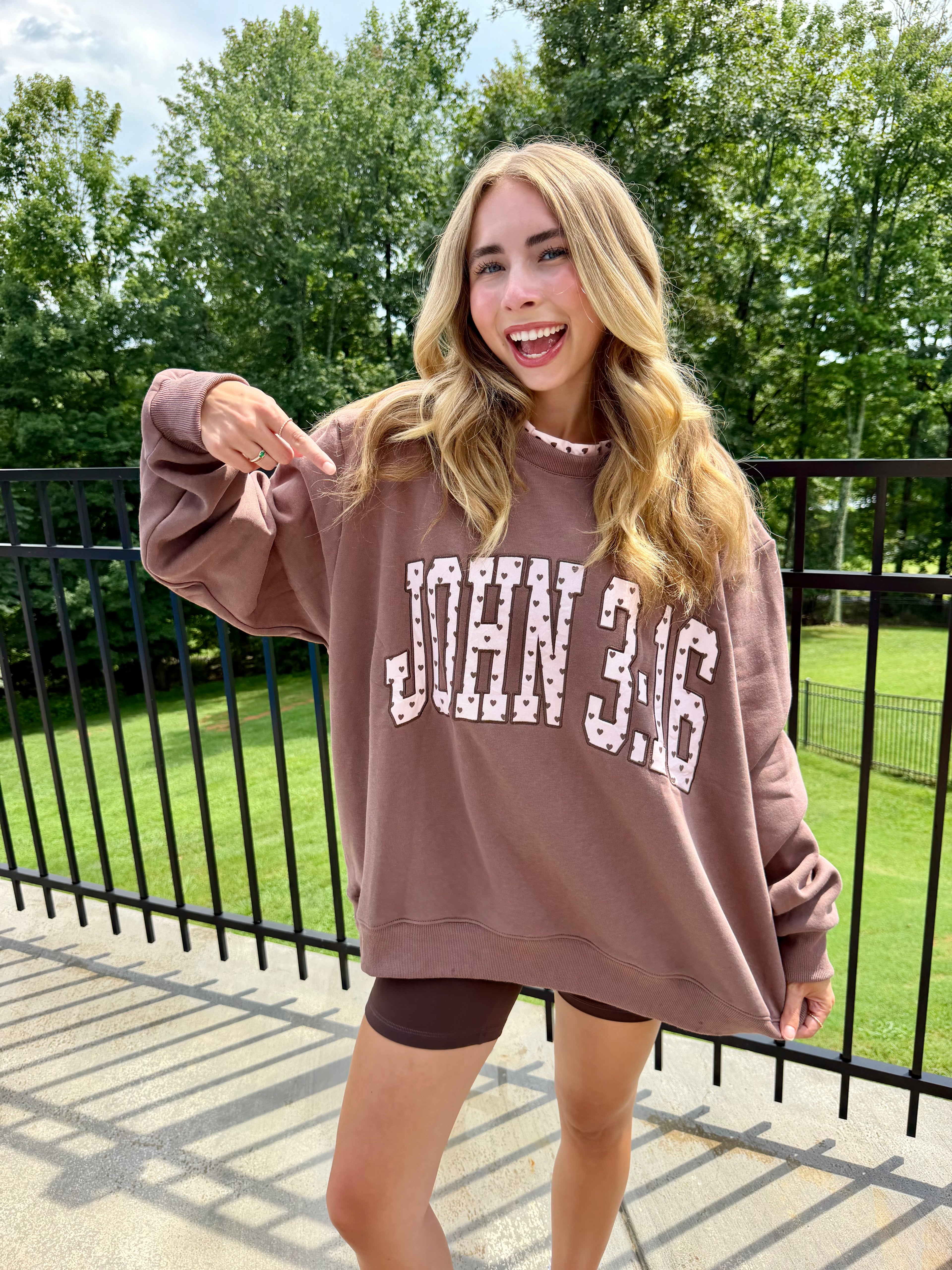 A woman wearing an oversized brown sweatshirt with 'JOHN 3:16' printed in white across the front, standing outdoors on a patio with trees and a fence in the background.