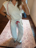 A woman modeling a light heather gray two-piece loungewear set with red piping and a red drawstring, standing on a patterned rug in a home setting.
