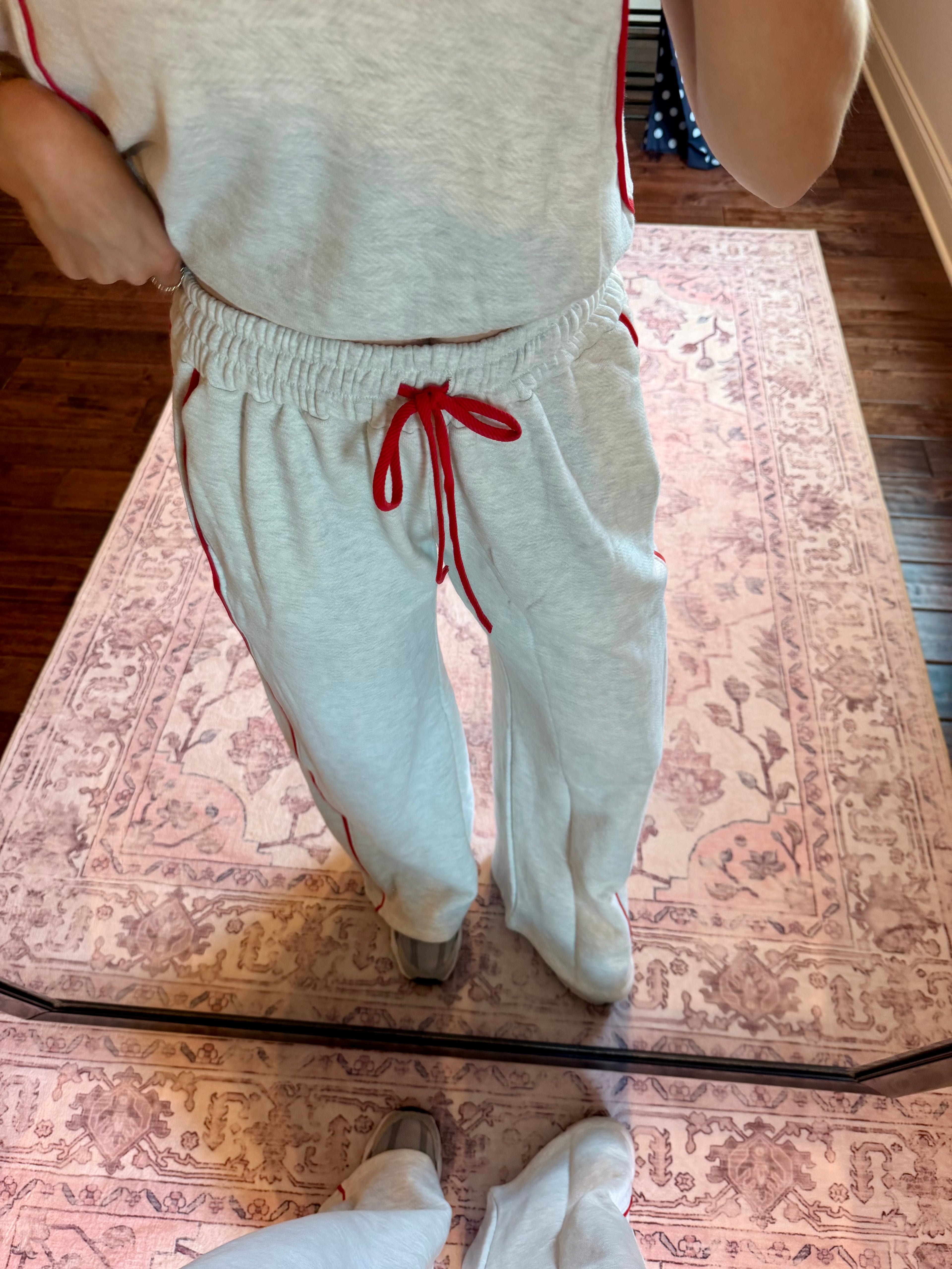 A person wearing light heather gray sweatpants with red side stripes and a red drawstring, standing on a patterned rug in front of a mirror.